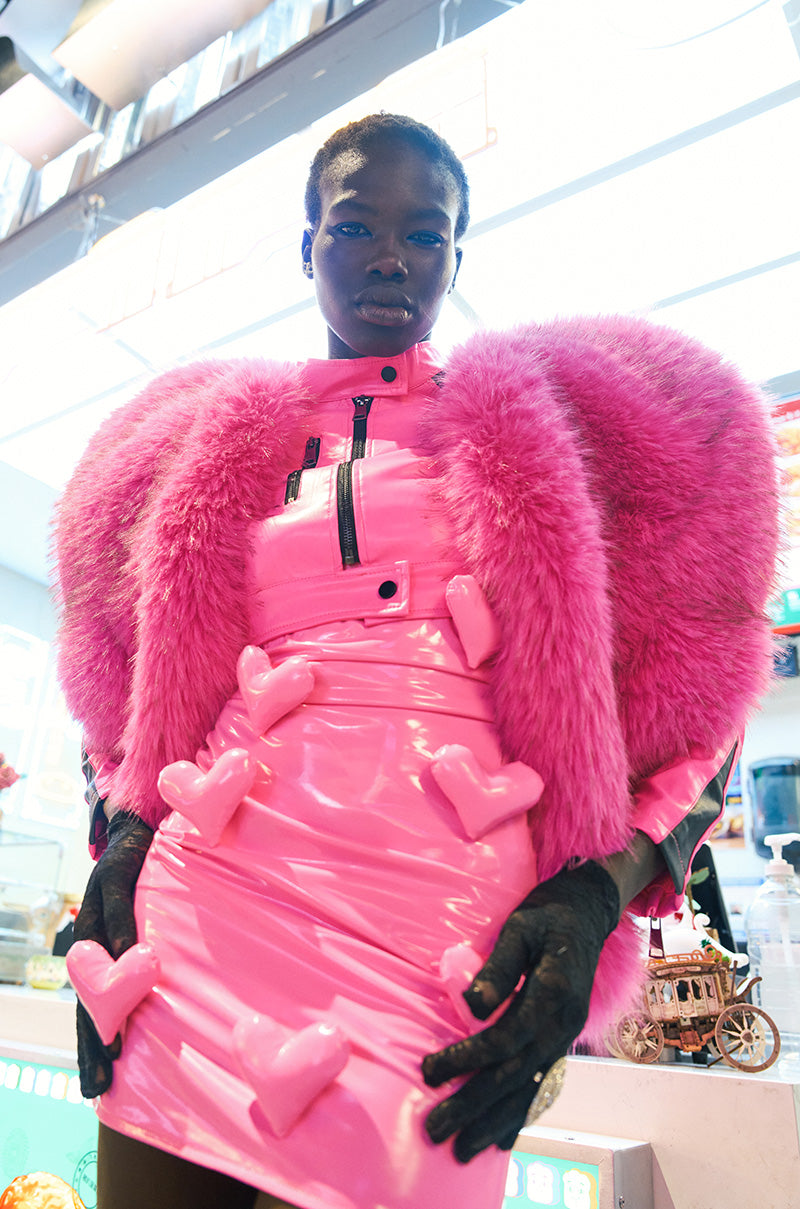 A person is showcasing the vibrant BARBIE DREAMS FAUX LEATHER MINI DRESS IN PINK, complete with a fluffy jacket and shimmering 3D heart appliques that capture attention. The striking ensemble is accentuated by black gloves, emphasizing the dress's bodycon fit, all within a bright and colorful indoor setting.