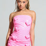 A person with braided hair wears the Barbie Dreams Faux Leather Mini Dress in pink, a strapless bodycon fit adorned with 3D heart shapes, paired with matching pink thigh-high stockings against a plain gray background.