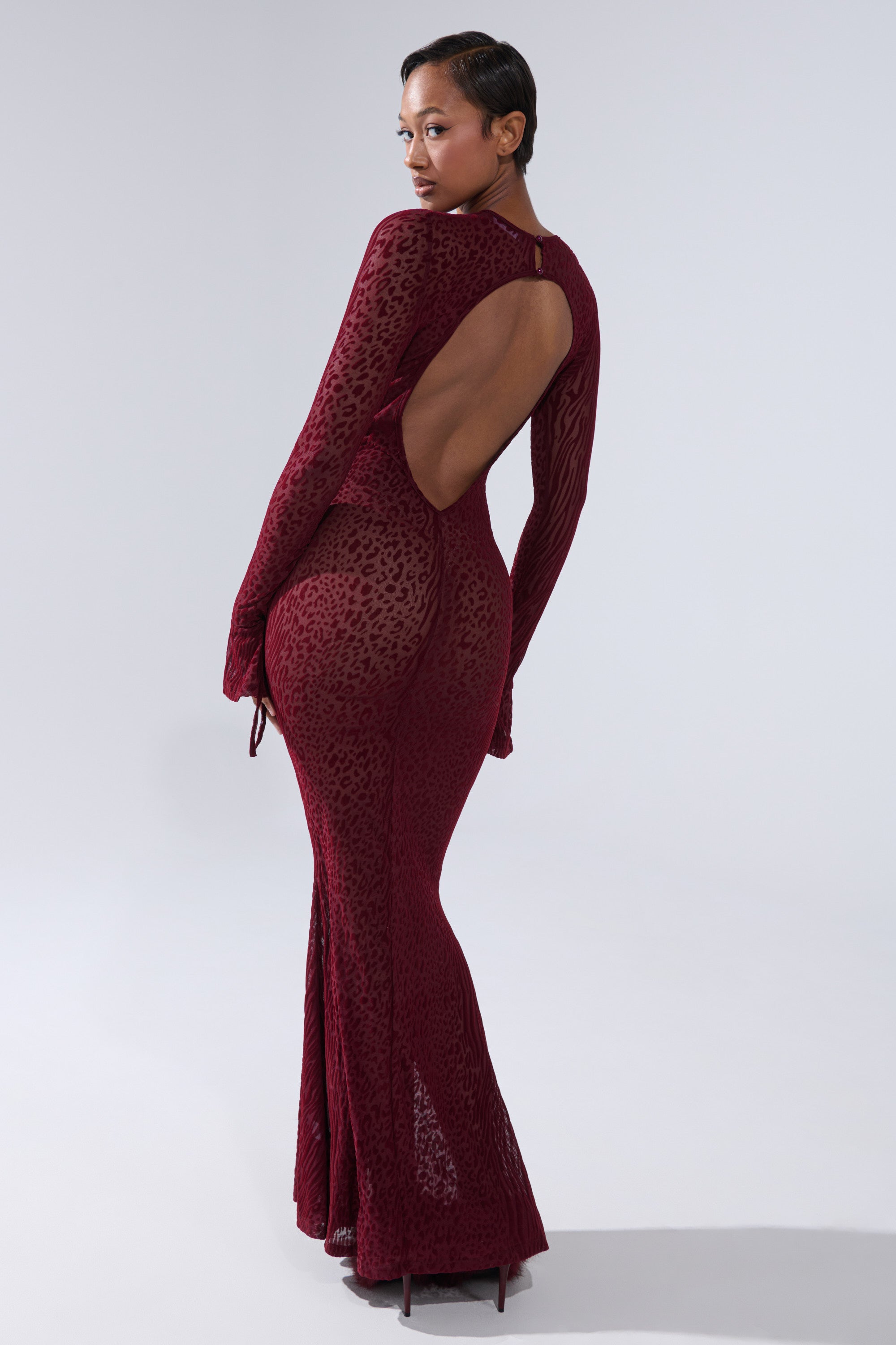 A person poses on a light gray background in the MY GOOD SIDE LEOPARD MESH MAXI DRESS IN BURGUNDY, featuring a fitted silhouette, long sleeves, animal print burnout design, open back, and looks over one shoulder.