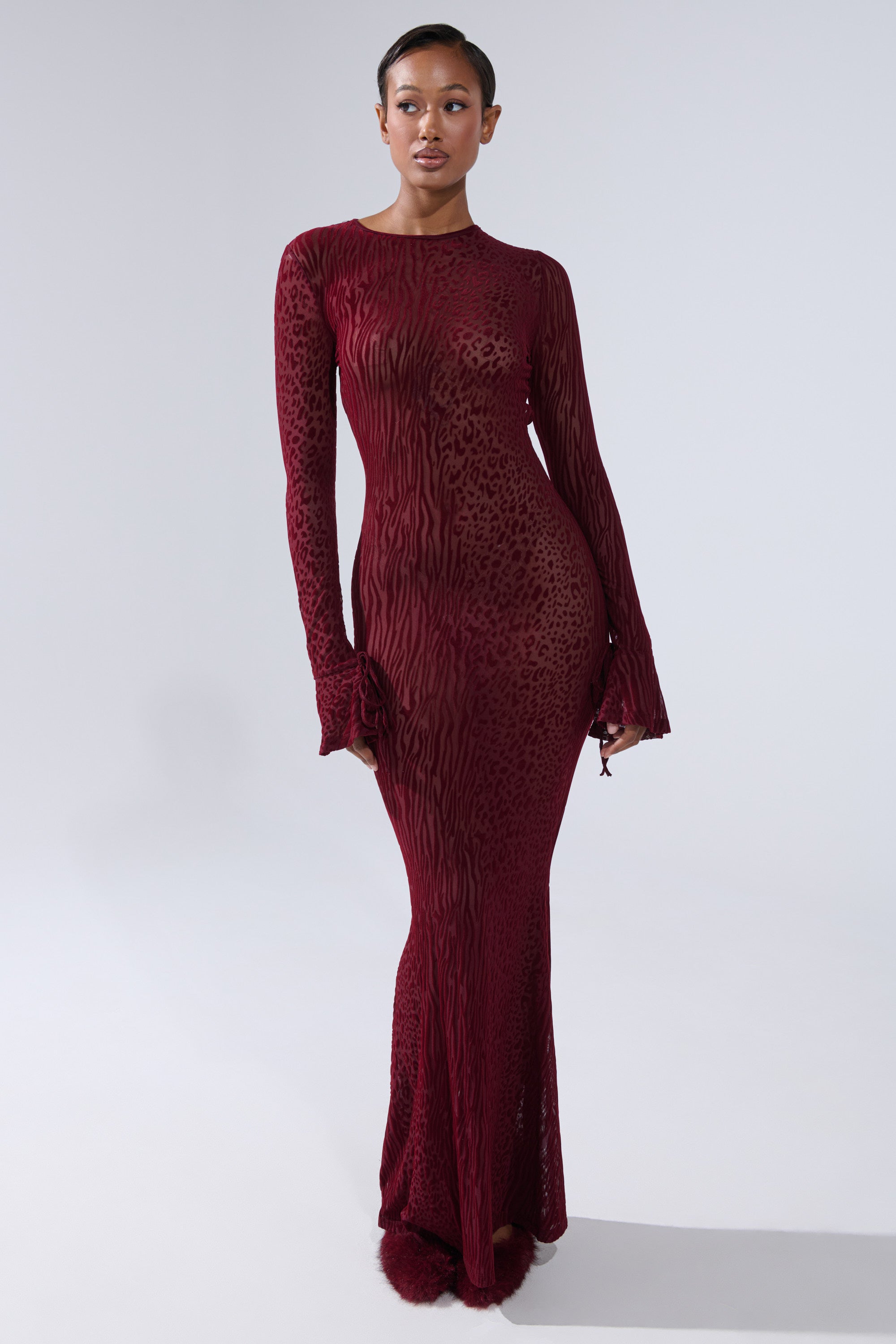 A woman poses confidently in the MY GOOD SIDE Leopard Mesh Maxi Dress in Burgundy, featuring long sleeves, a fitted silhouette with a subtle flare at the hem, and an animal print burnout pattern against a plain background.