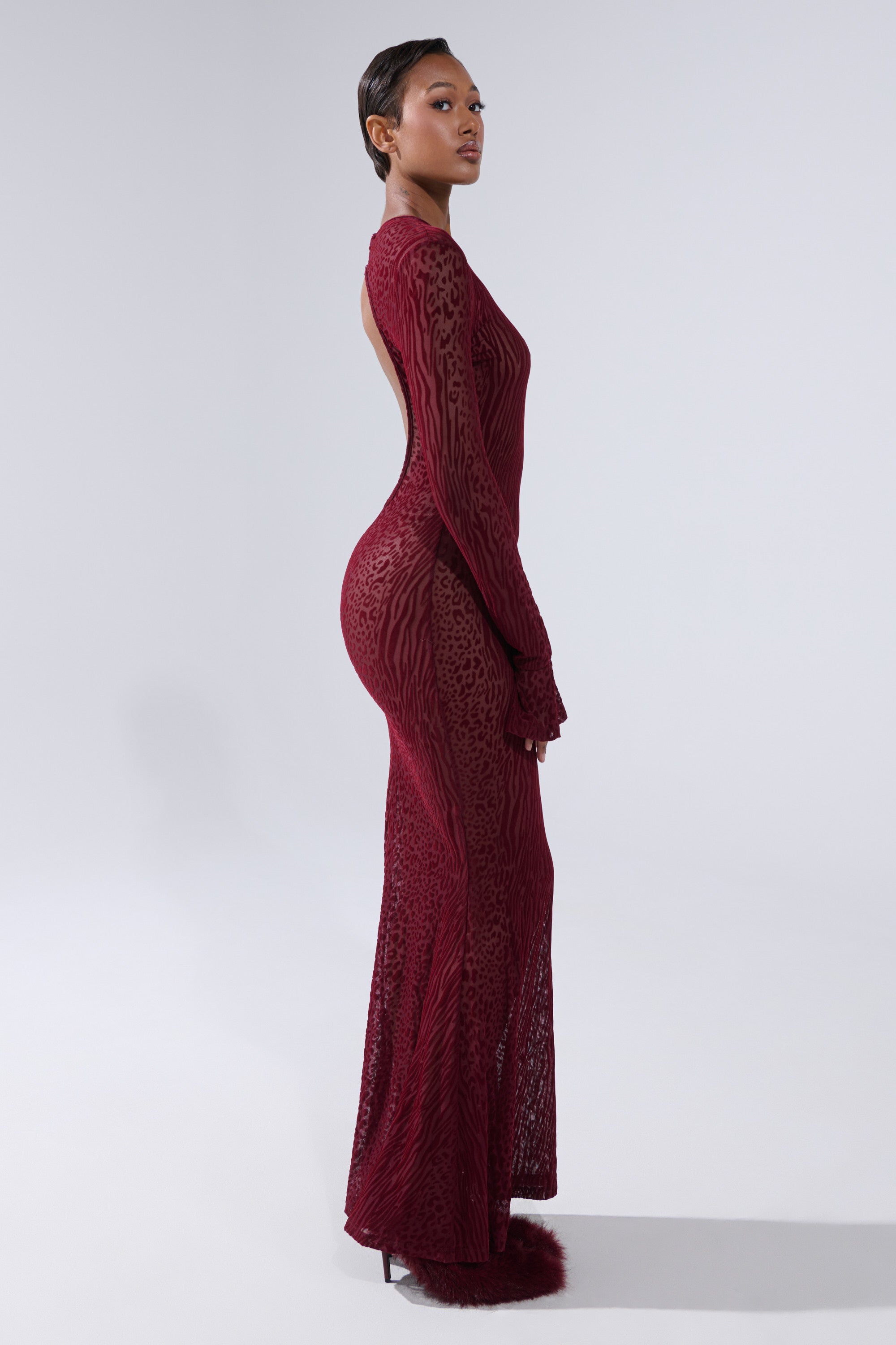 A woman with short hair stands in profile against a plain background, wearing the MY GOOD SIDE Leopard Mesh Maxi Dress in Burgundy, a long, fitted dress with textured patterns and long sleeves, looking slightly upwards.