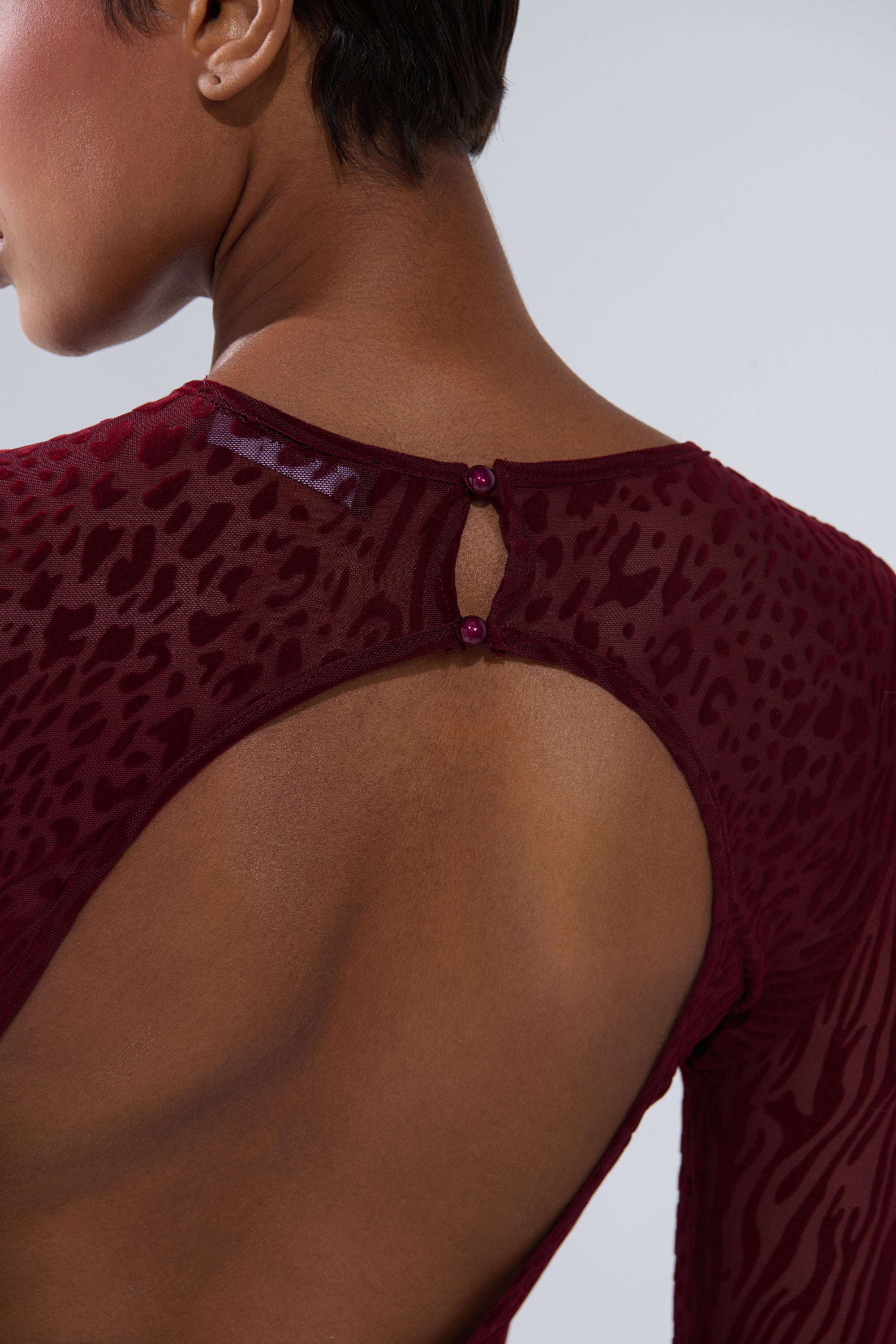 A close-up shows a person wearing the MY GOOD SIDE Leopard Mesh Maxi Dress in Burgundy, featuring a sheer animal print, an open back with two button closures at the neckline, and their short hair and neck visible.