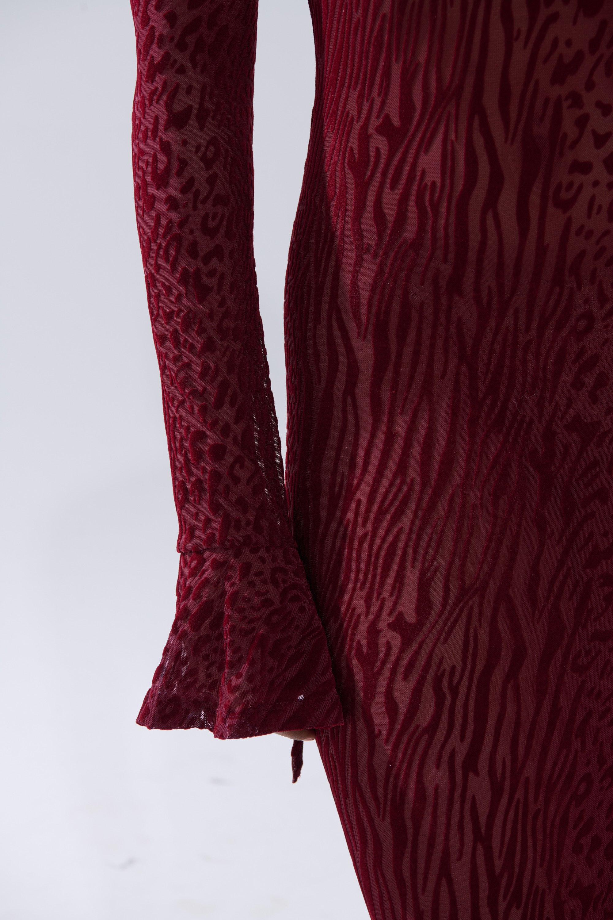 Close-up of a person wearing the MY GOOD SIDE LEOPARD MESH MAXI DRESS IN BURGUNDY, a long-sleeved, form-fitting maxi with an animal print burnout texture. The plain background highlights the sleeve and side details of this striking dress.