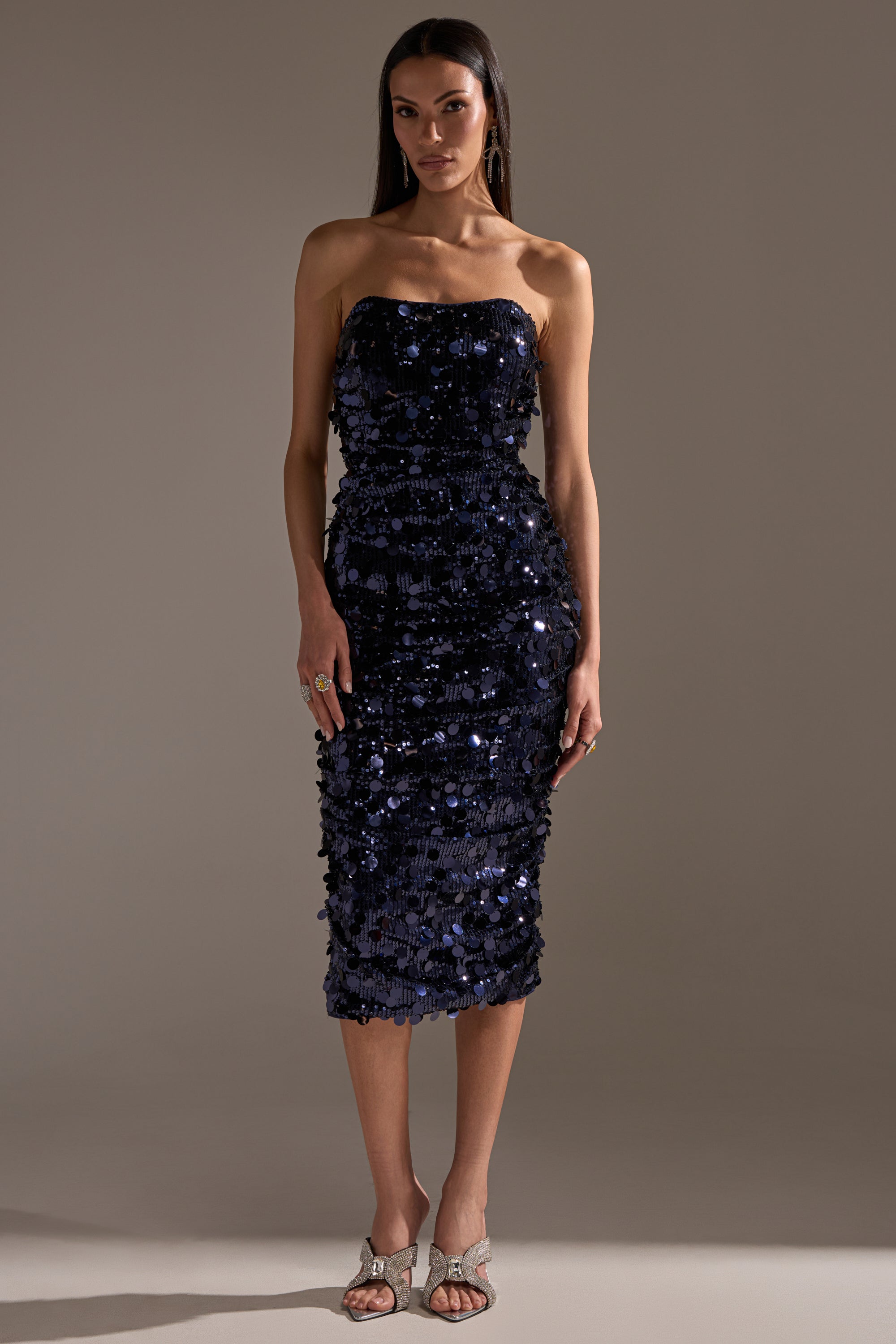 A woman poses against a plain backdrop wearing the WHO'S THAT GIRL SEQUIN MIDI DRESS—a fitted, navy, textured sequin strapless dress. She has long dark hair and accessorizes with earrings, rings, and open-toe heels.