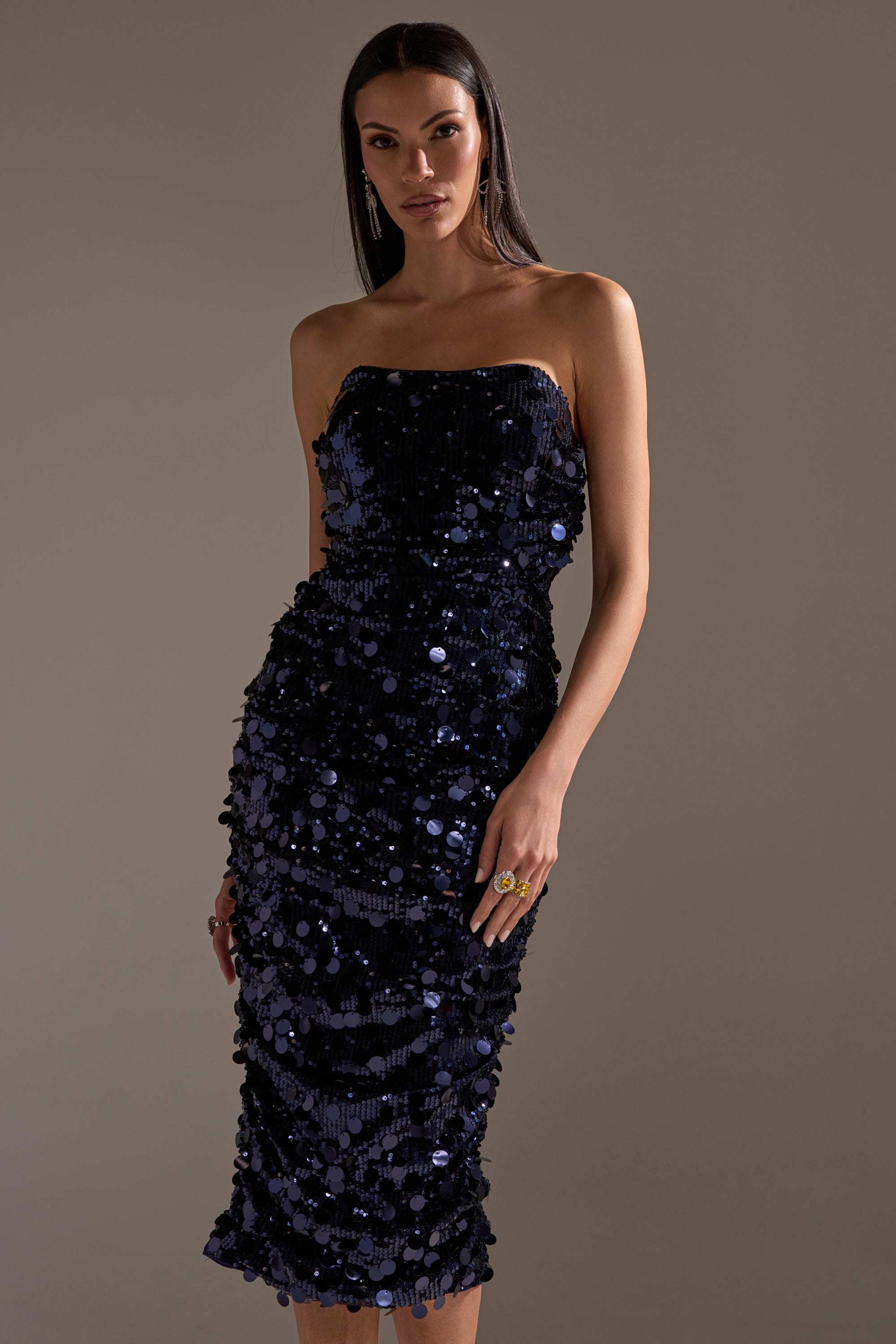 A woman with long, straight hair stands against a plain background wearing the WHO'S THAT GIRL SEQUIN MIDI DRESS—a strapless, navy sequin dress falling below the knee—styled with earrings and a statement ring.