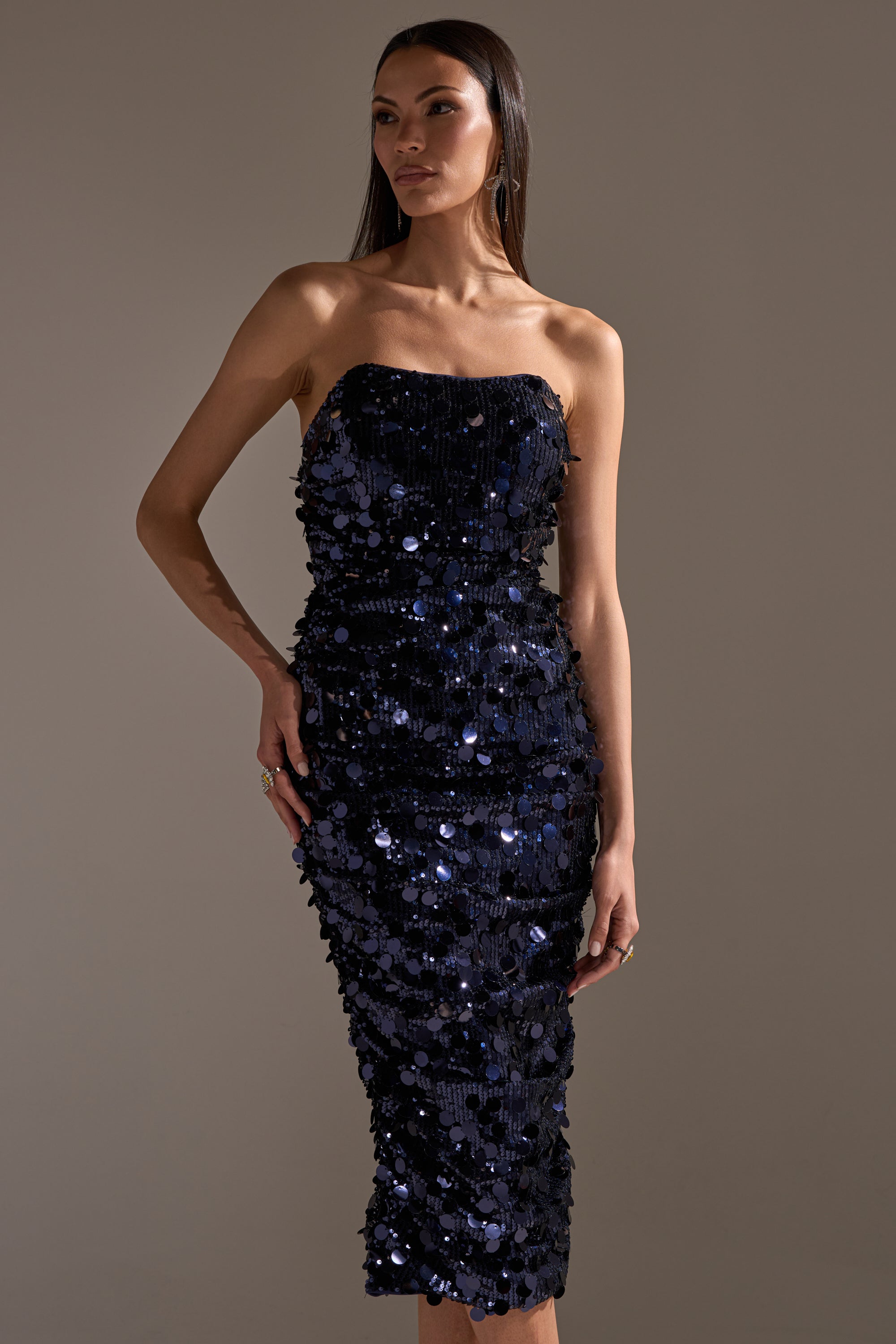 A woman stands against a plain background wearing the WHO'S THAT GIRL SEQUIN MIDI DRESS—a form-fitting, navy sequin, strapless party dress that falls below the knee. She has long straight hair and relaxed arms at her sides.