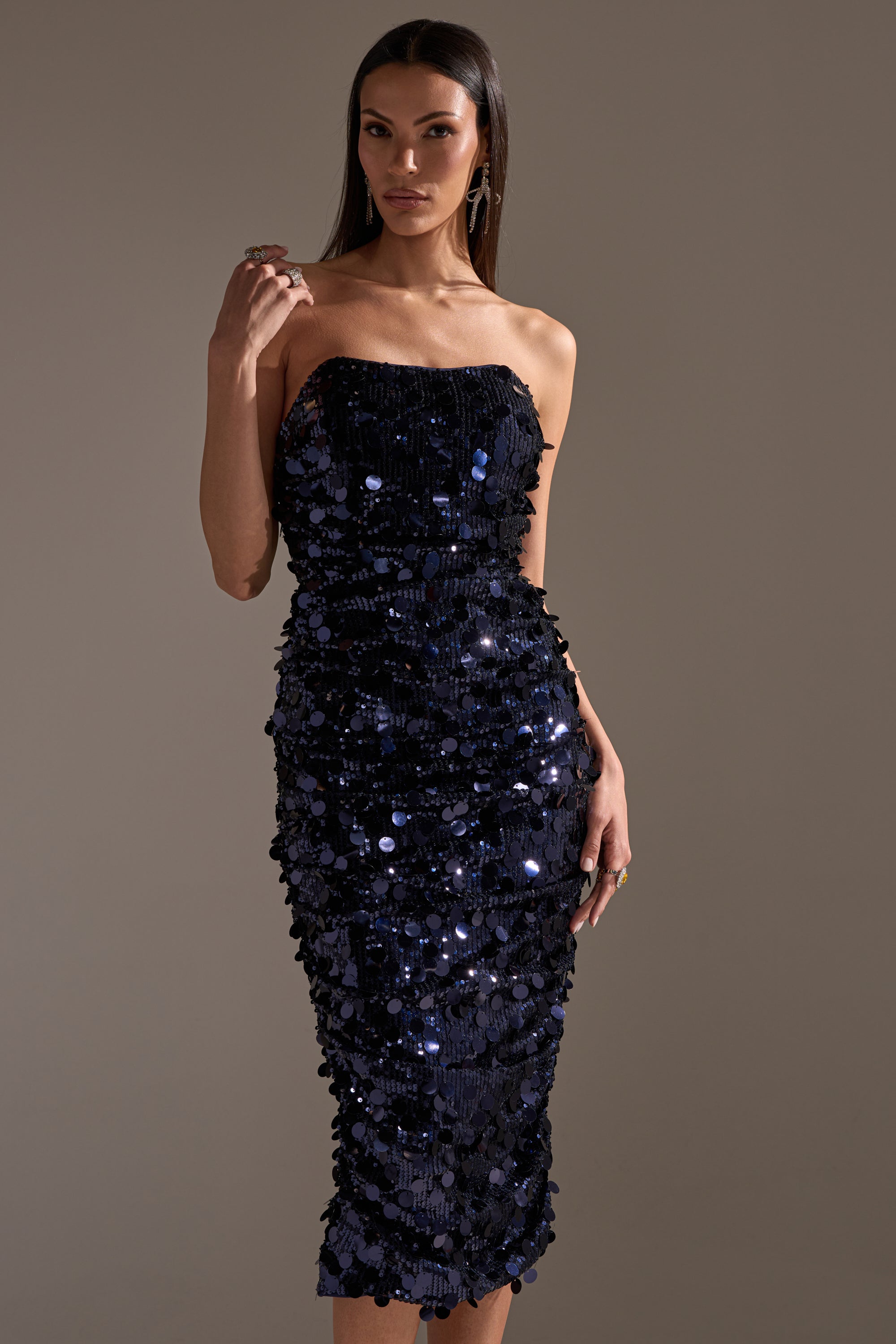 A woman stands confidently against a plain background, wearing the WHO'S THAT GIRL SEQUIN MIDI DRESS in strapless navy, with one hand raised near her shoulder as she gazes at the camera.