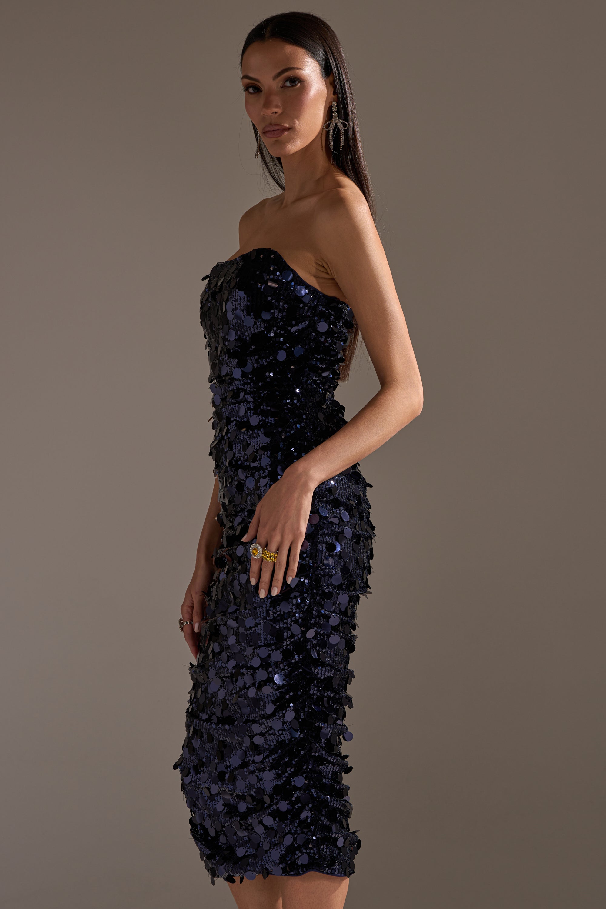 A woman models the WHO'S THAT GIRL SEQUIN MIDI DRESS—a form-fitting, strapless navy sequin dress falling below the knee. She has long straight hair, large hoop earrings, and multiple rings, set against a plain neutral background.