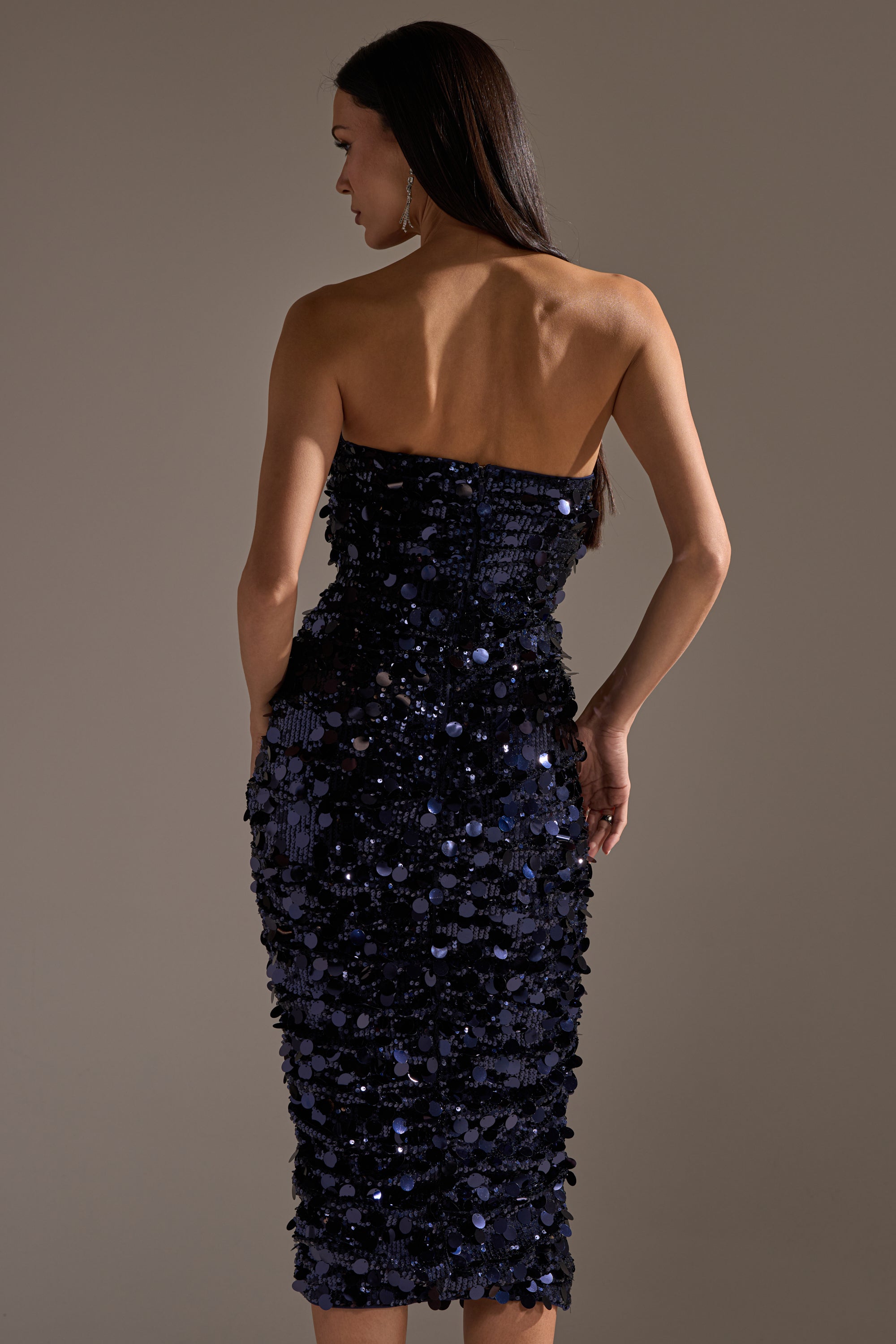 A woman with long dark hair faces away, wearing the WHO'S THAT GIRL SEQUIN MIDI DRESS—a strapless, navy, form-fitting sequin midi dress that falls below the knees and sparkles against a softly lit background.