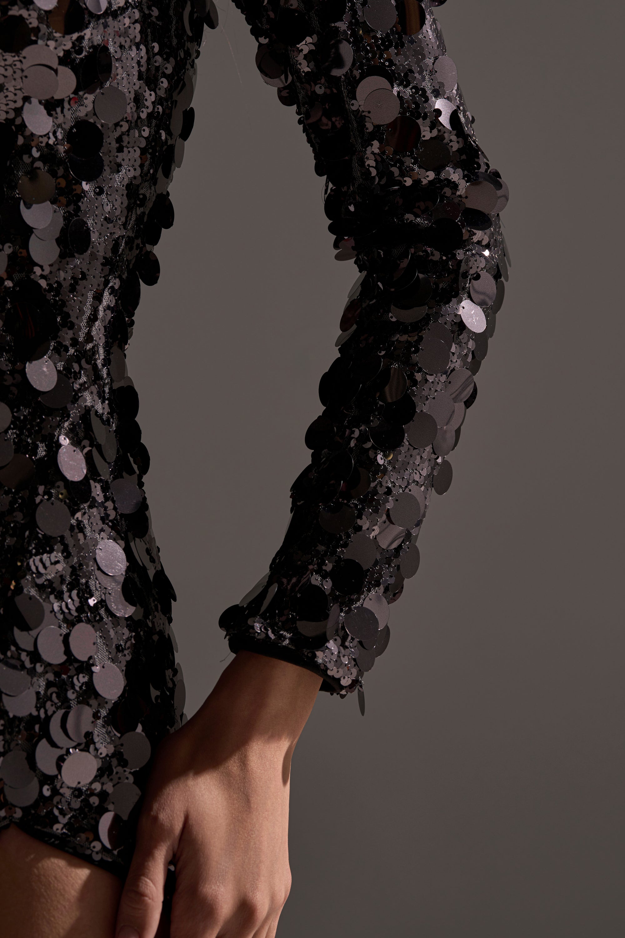A close-up of a person’s arm and hand wearing the DINNER AND A SHOW SEQUIN MAXI DRESS, a long-sleeved black sequin gown, against a plain gray background.