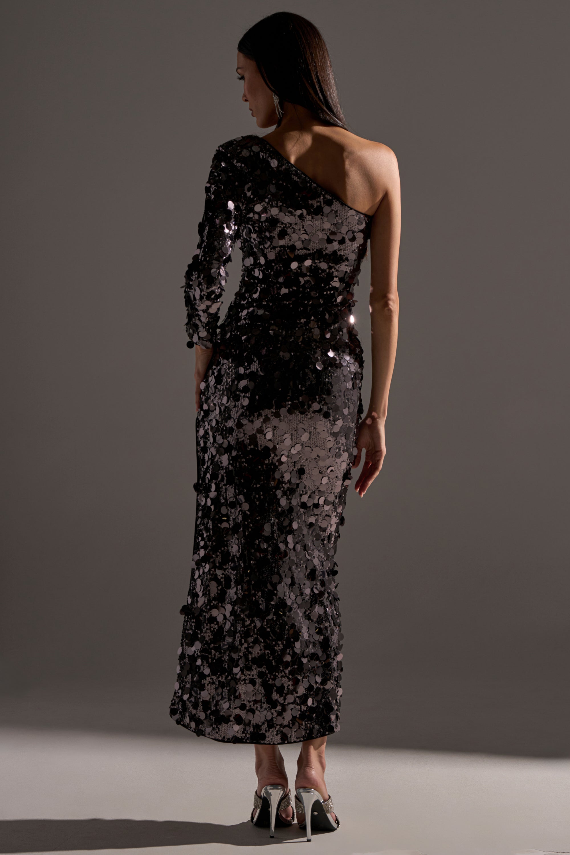 A woman stands with her back turned, wearing the DINNER AND A SHOW SEQUIN MAXI DRESS and black-and-white high heels in front of a plain gray background.