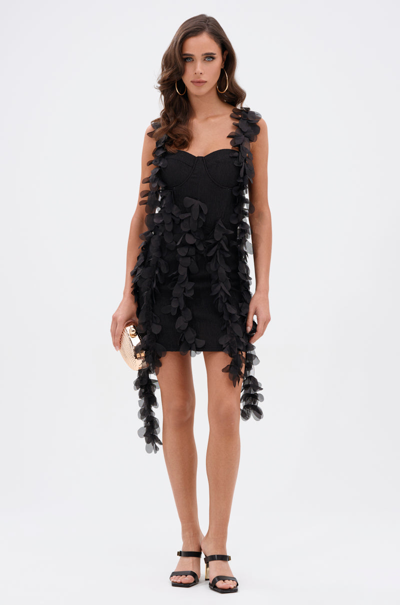 A woman stands against a white background wearing the COVER GIRL MINI DRESS WITH FLORAL APPLIQUES, paired with black sandals, gold hoop earrings, and holding a small gold clutch.