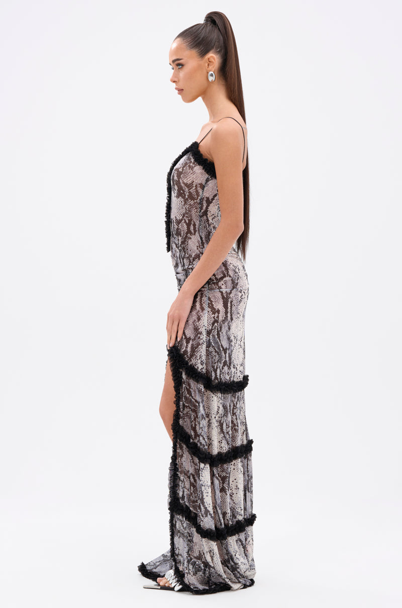 Woman in profile wears the I BITE BACK PRINTED MAXI DRESS with a plunging V-neckline and black fringe trim, paired with large earrings and a sleek high ponytail, against a plain white background.