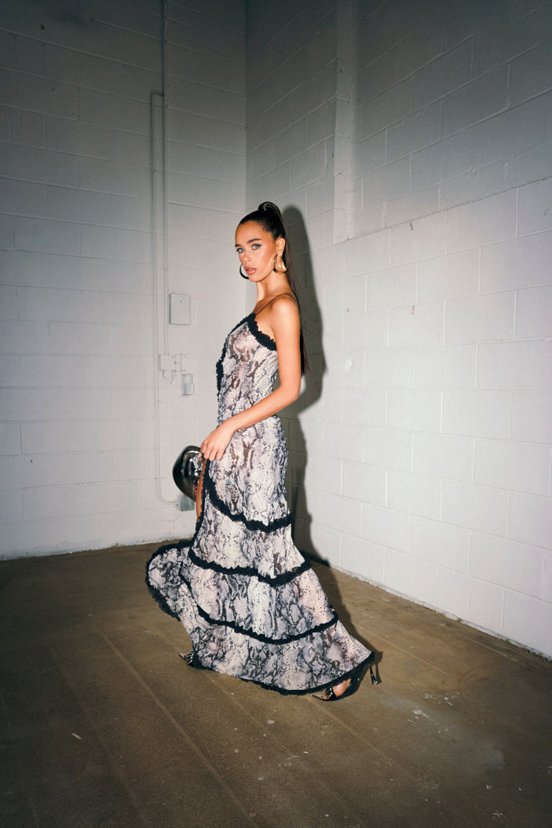 A woman with a high ponytail wears the I BITE BACK PRINTED MAXI DRESS, featuring a sleeveless design, lace pattern, black trim, ruffles, and a plunging V-neckline, standing in a plain room with white brick walls and concrete floor.