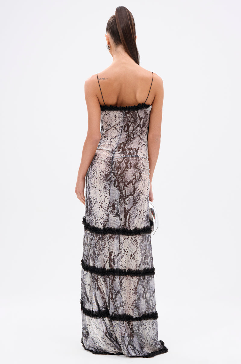 A woman faces away, wearing the I BITE BACK PRINTED MAXI DRESS—a floor-length snakeskin print mesh maxi with thin straps and black fringe rows. She holds a silver clutch and has long, straight hair styled in a high ponytail.
