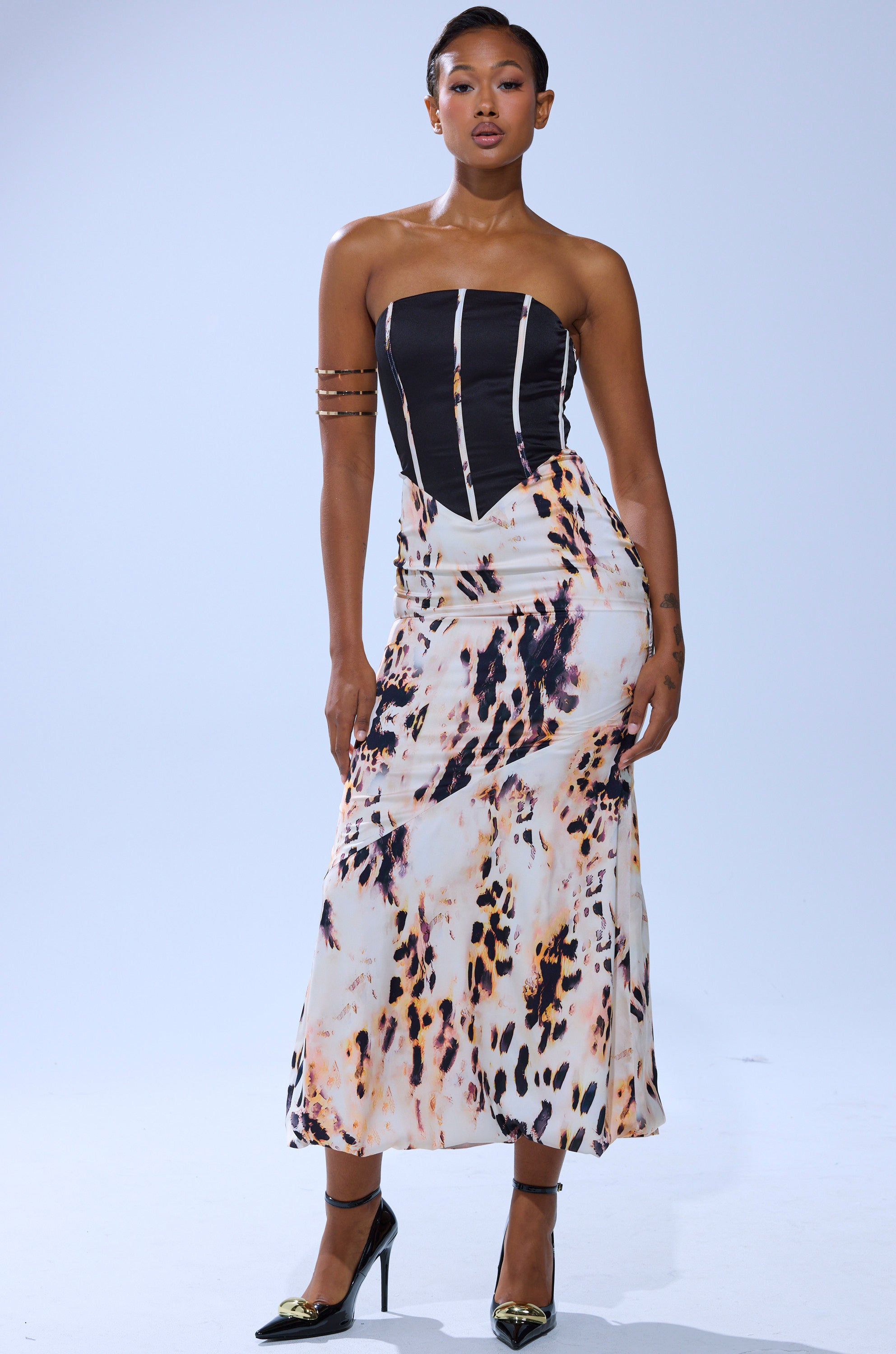 A woman models THE REAL DEAL PRINTED MAXI DRESS, featuring a strapless black corset-style top with white boning and a high-waisted animal print skirt, accessorized with pointed black heels and gold arm jewelry against a simple light background.