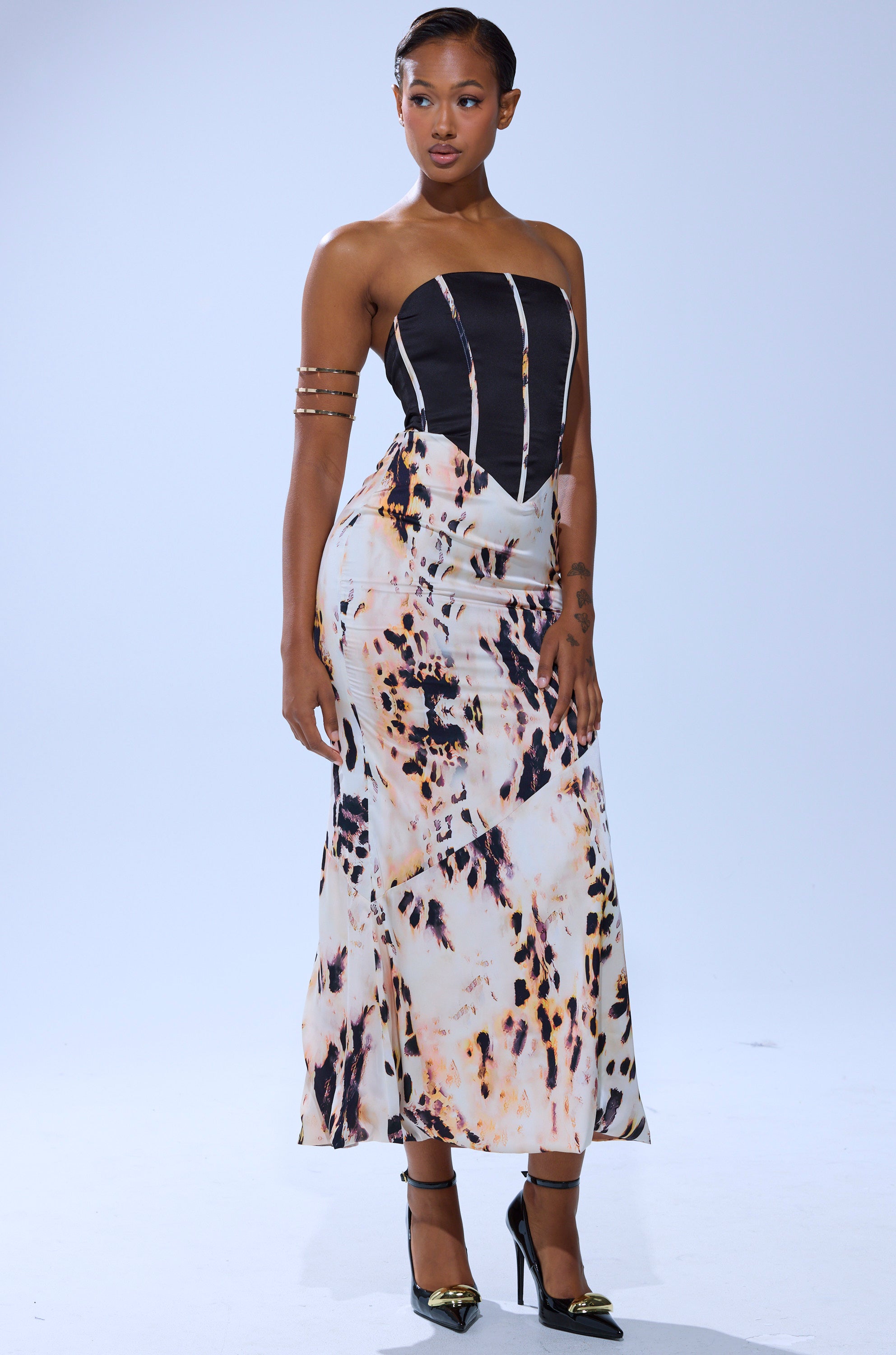 A woman models THE REAL DEAL PRINTED MAXI DRESS, a strapless black and animal-print maxi with a long skirt. She pairs it with black heels featuring gold toes, gold arm cuffs, and has short slicked-back hair against a light gray background.