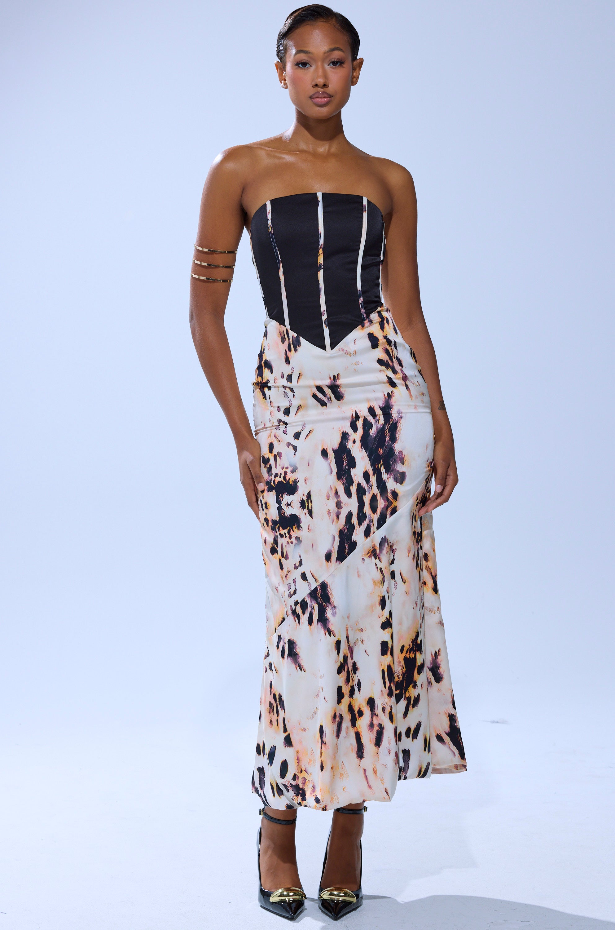 A woman stands confidently against a plain light background in THE REAL DEAL PRINTED MAXI DRESS, featuring a strapless black corset-style top and long, fitted abstract print skirt. She completes the look with black pointed heels and gold arm cuffs.