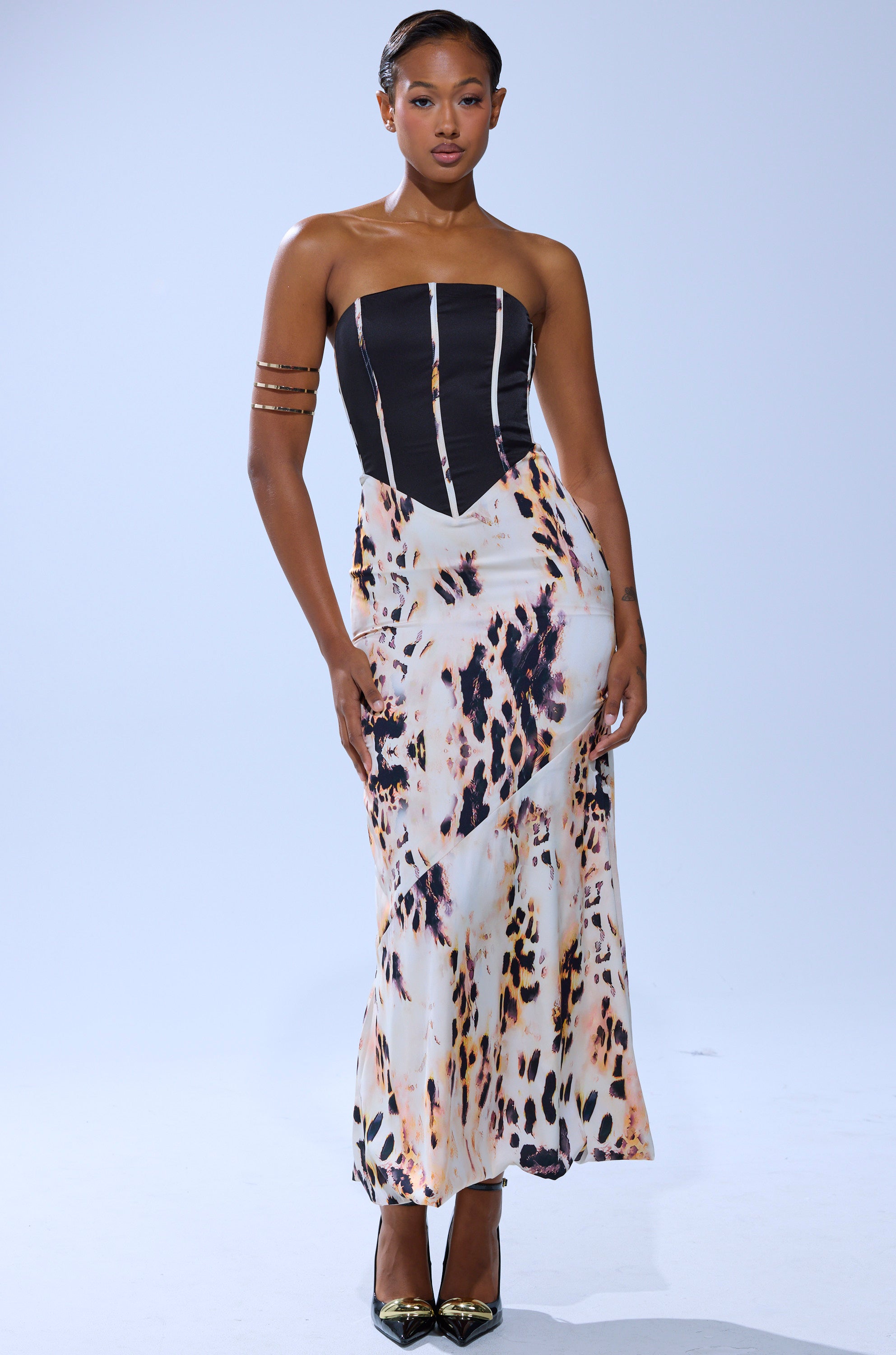 A woman models THE REAL DEAL PRINTED MAXI DRESS, featuring a black corset-style satin top and flowing animal-print maxi skirt. She pairs it with metallic heels, a gold arm cuff, slicked-back hair, and stands before a plain light background.