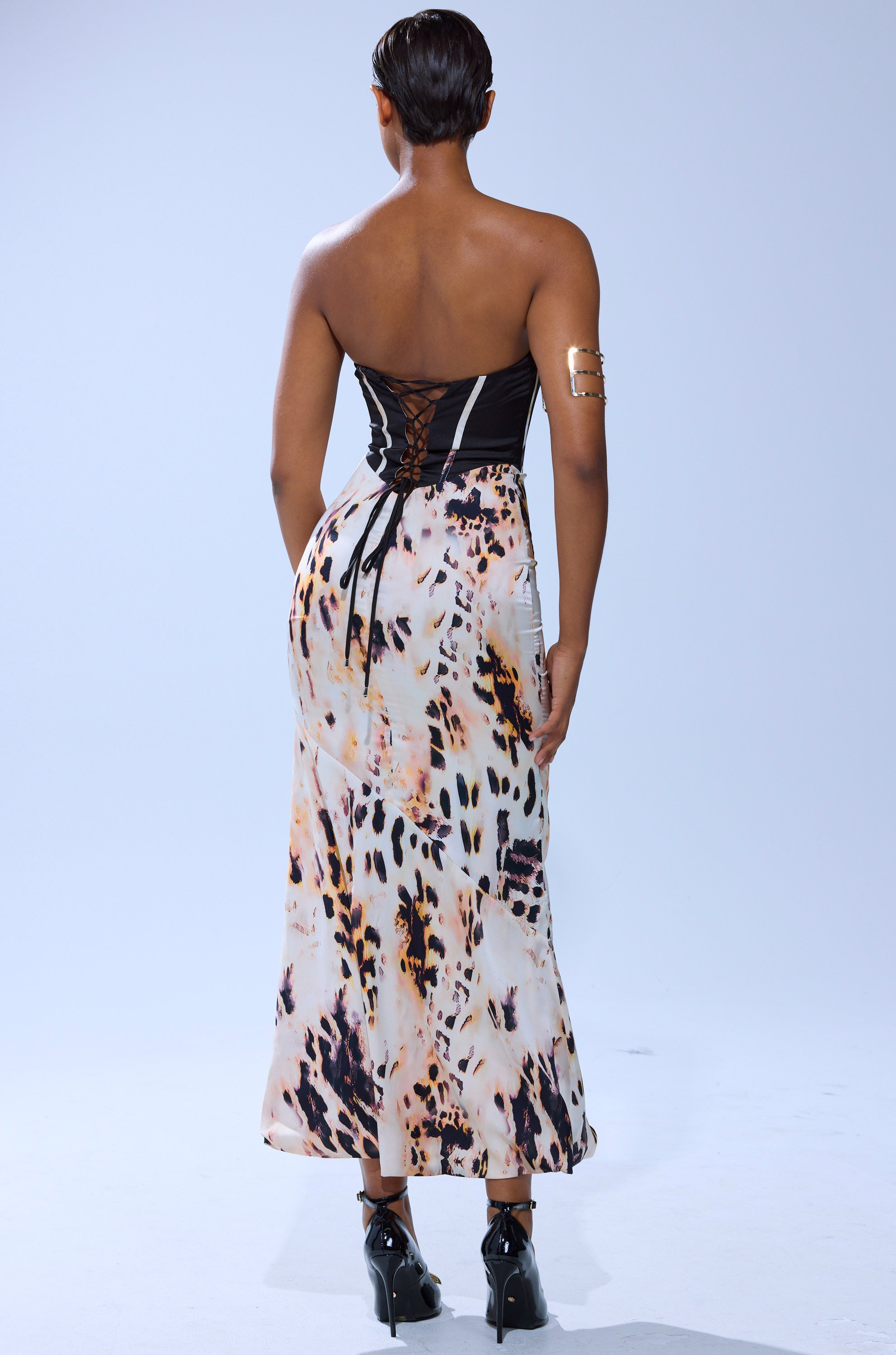 A woman with short hair wears THE REAL DEAL PRINTED MAXI DRESS over a black corset top, styled with a gold arm cuff and black heeled shoes, standing against a plain light background.