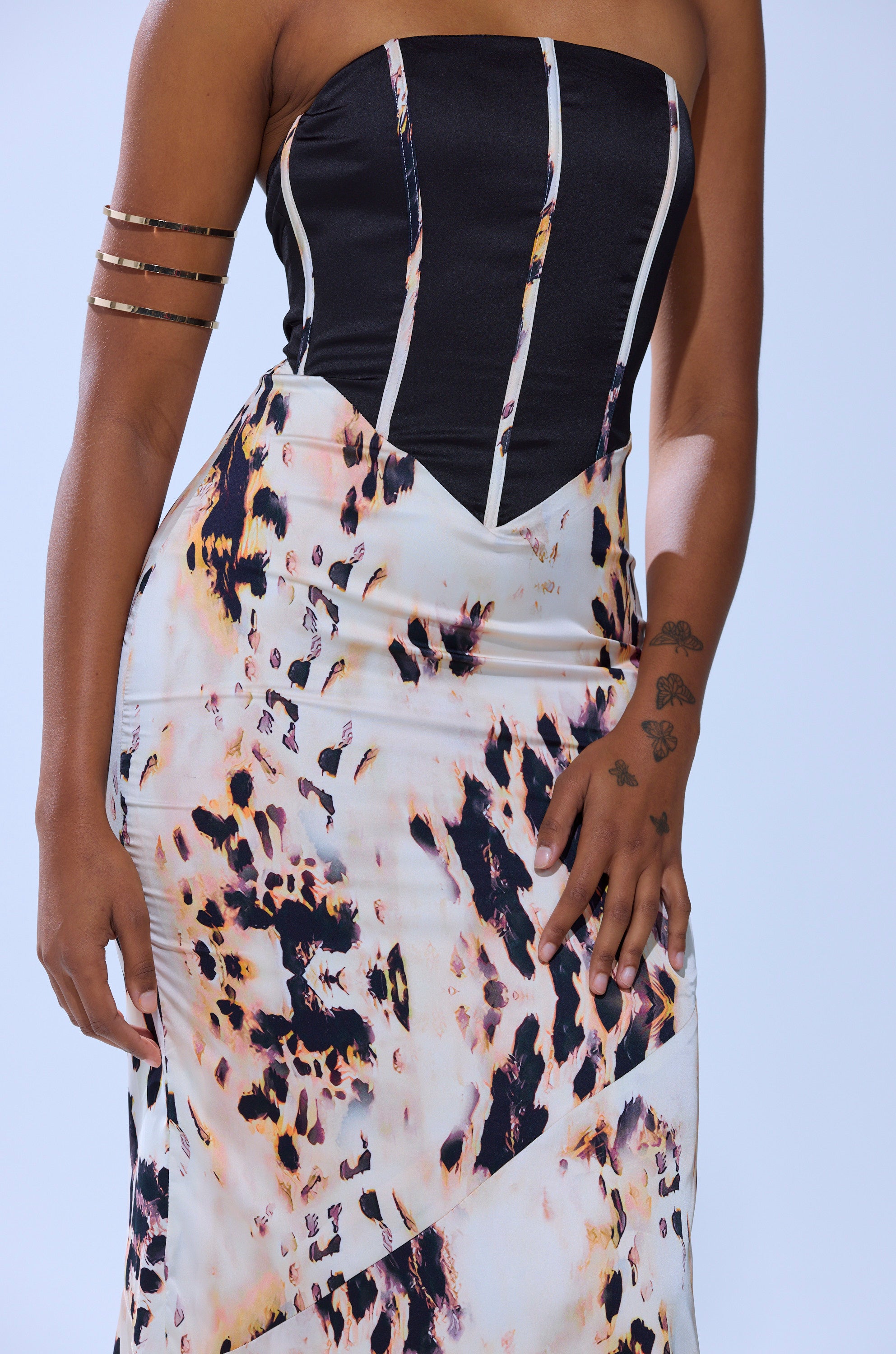 Wearing THE REAL DEAL PRINTED MAXI DRESS—a strapless black and white animal-print sheath—paired with gold arm cuffs and showcasing small tattoos on their hand and wrist, the person stands against a light background.