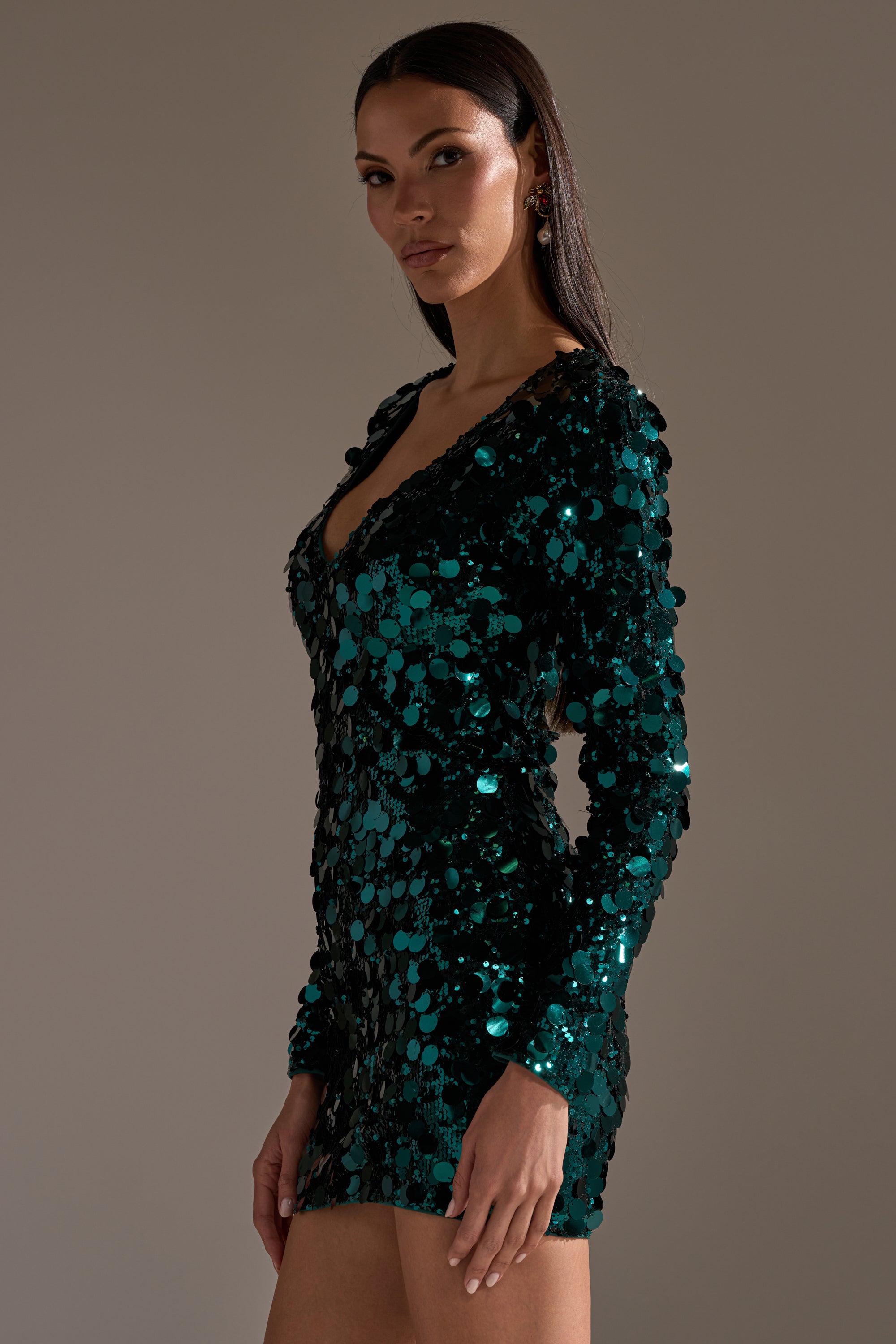 A woman models the DANCING QUEEN SEQUIN MINI DRESS, a long-sleeved, deep green sequin mini dress with a deep V-neckline. She stands against a plain background, confidently looking at the camera with straight dark hair.