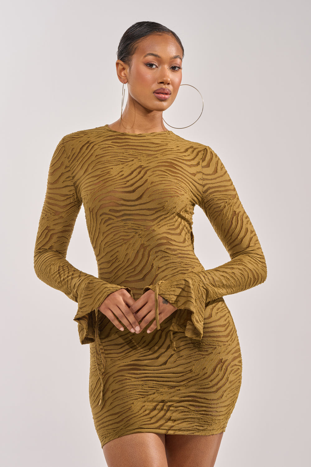 A woman poses confidently in the SEE ME WIN MESH MINI DRESS IN OLIVE, a long sleeve mini dress featuring textured fabric. She accessorizes with large hoop earrings and has her hair pulled back. 1
