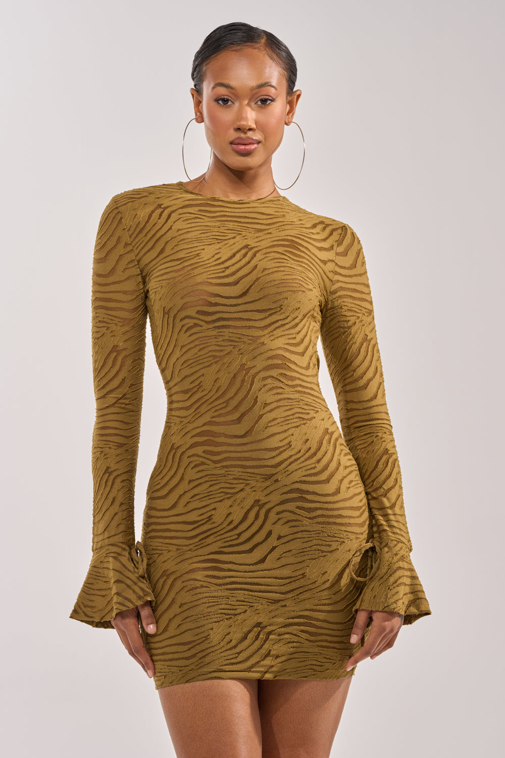A woman poses confidently against a plain, light background wearing the SEE ME WIN Mesh Mini Dress in Olive, featuring a fitted silhouette, long sleeves, and flared cuffs. 2