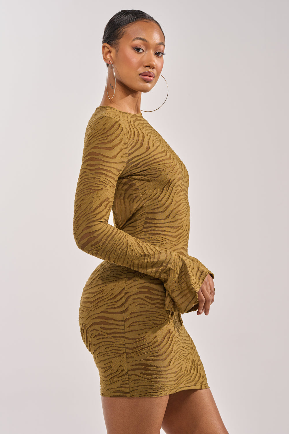 A woman poses sideways in the SEE ME WIN Mesh Mini Dress in olive, a fitted long sleeve mini dress with a striking olive and brown tiger-stripe pattern, paired with large hoop earrings and hair pulled back against a plain light background. 3