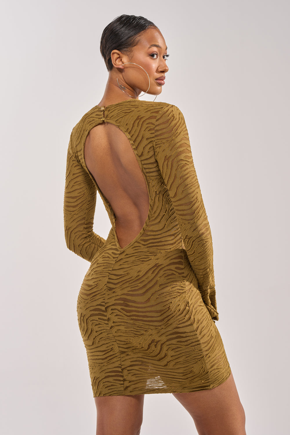 A woman stands against a plain background, looking over her shoulder in the SEE ME WIN Mesh Mini Dress in Olive—a form-fitting, long sleeve dress with a zebra print and open back. 4