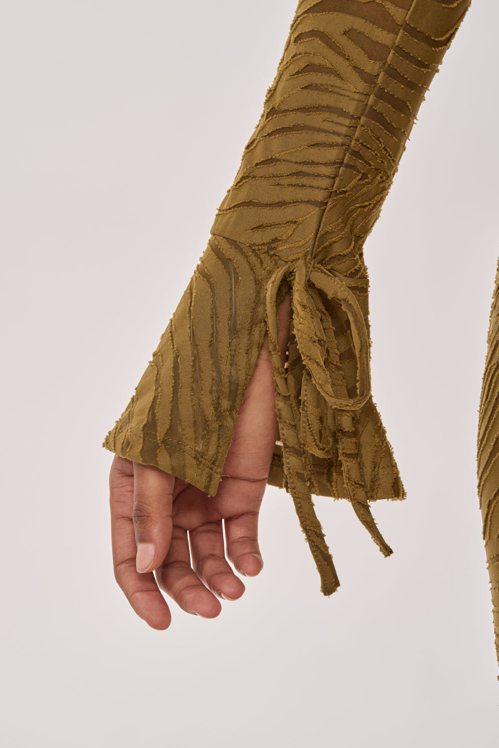 A close-up of a hand and lower arm in a tiger-striped olive green sleeve with flared cuff and tie detail from the SEE ME WIN MESH MINI DRESS IN OLIVE, against a plain light gray background. 7