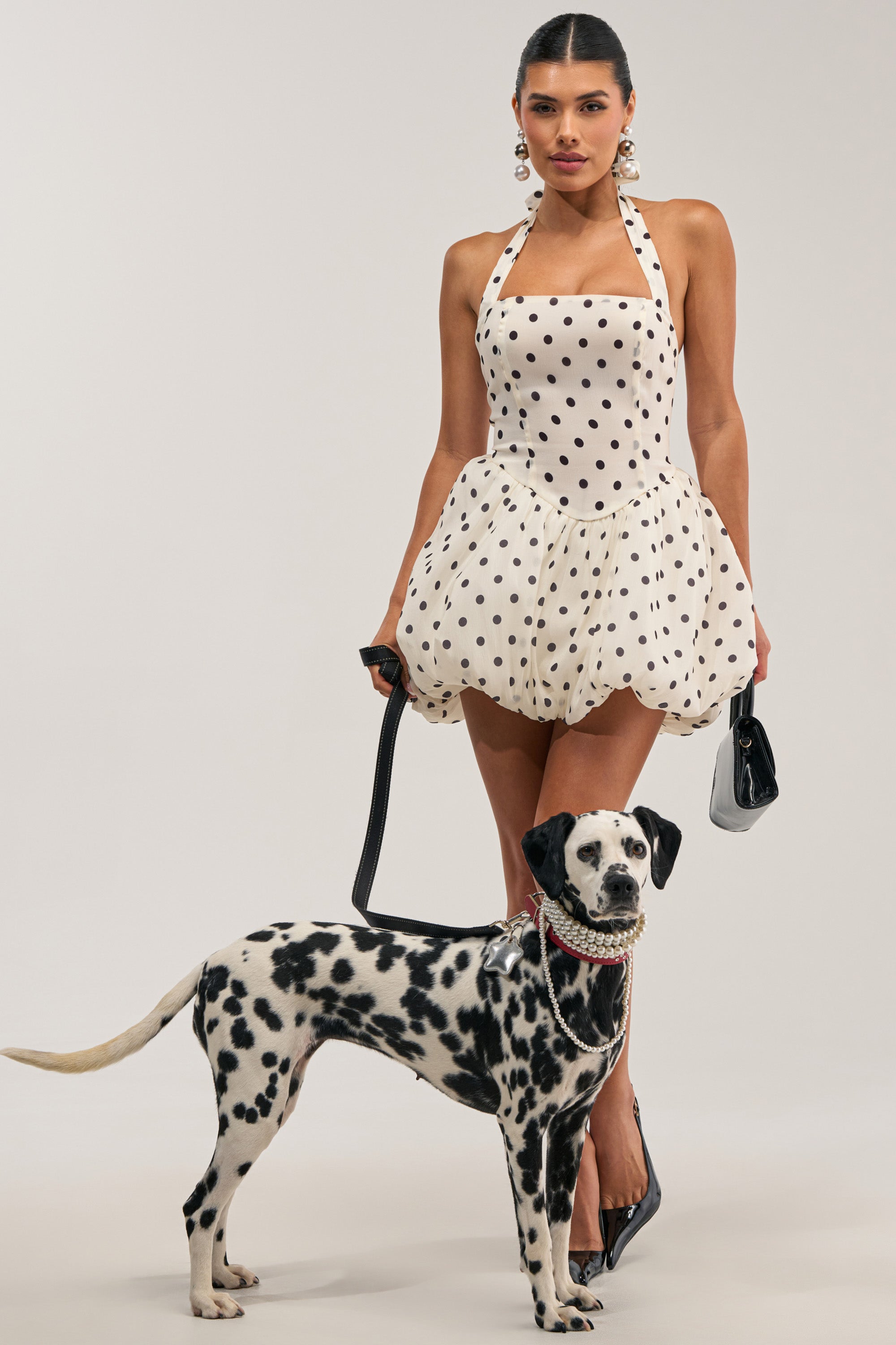 A woman wearing the FELT CUTE POLKA DOT MINI DRESS—a cream-colored mini dress with polka dots and a bubble hem—pairs her look with pearl earrings as she stands holding a black leash attached to a Dalmatian dog. The dog, styled in a pearl collar and necklace, poses with her against a plain light gray background.