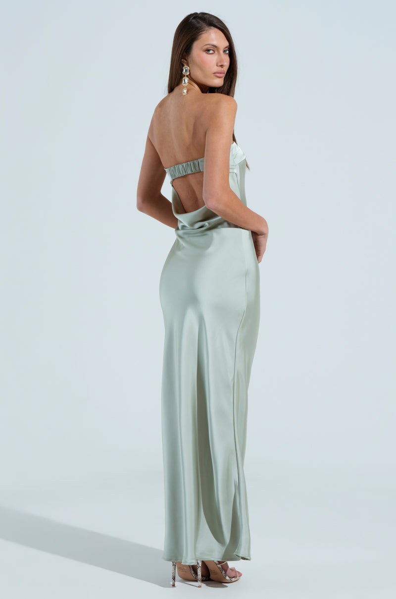 A woman in THE ONE THAT GOT AWAY SATIN MIDI DRESS, a strapless light blue mermaid silhouette with a cut-out back, faces away from the camera. She wears dangling earrings and silver heels, elegantly posing against a plain light background.