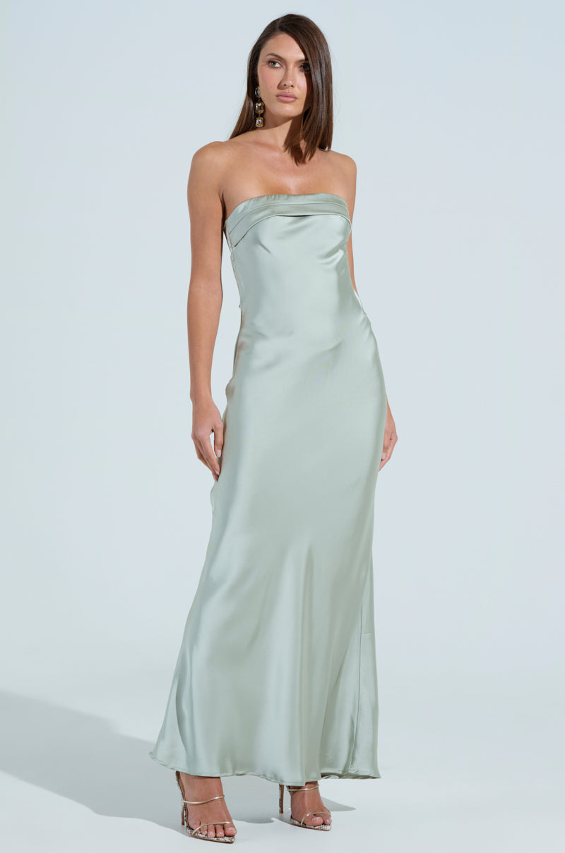 A woman stands against a plain backdrop in "THE ONE THAT GOT AWAY SATIN MIDI DRESS," showcasing its silvery-blue hue with a strapless, mermaid silhouette. She accessorizes with drop earrings and metallic strappy heels, as her straight hair flows over her shoulders.