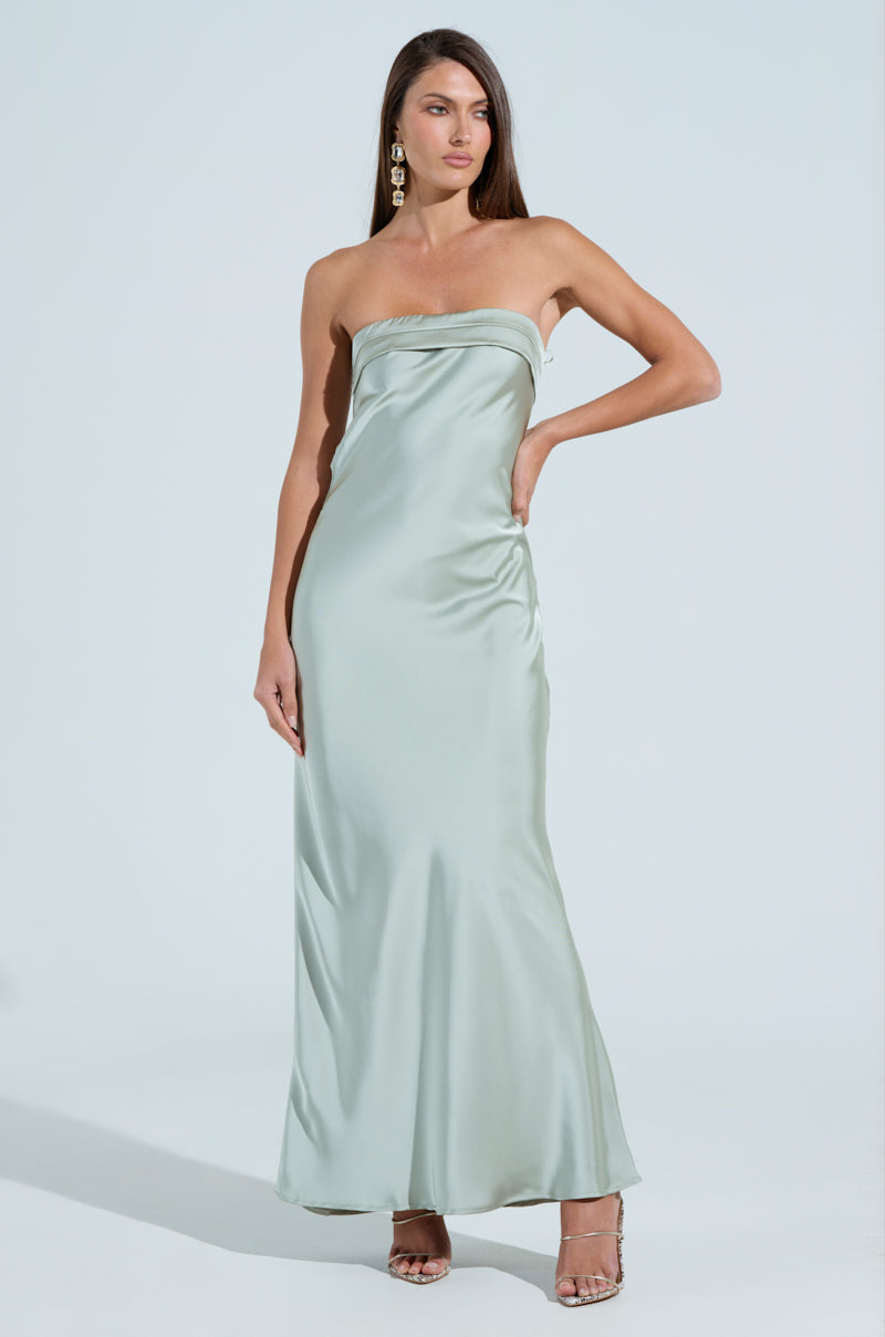 Wearing "The One That Got Away Satin Midi Dress," a person elegantly poses against a plain background. The strapless, light green ensemble complements their long hair, while high heels and large, dangling earrings add a touch of sophistication.