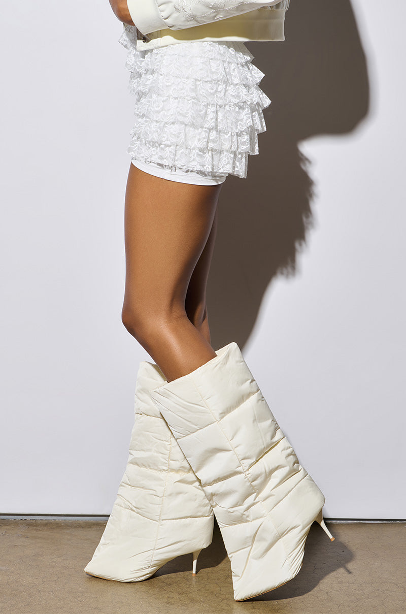 Displaying a unique sculptural silhouette, a person with tanned legs is wearing a white ruffled skirt and the DREAMSICLE CREAM PUFFER STILETTO BOOT, which features an oversized quilted upper. They stand on a concrete floor against a plain white background.