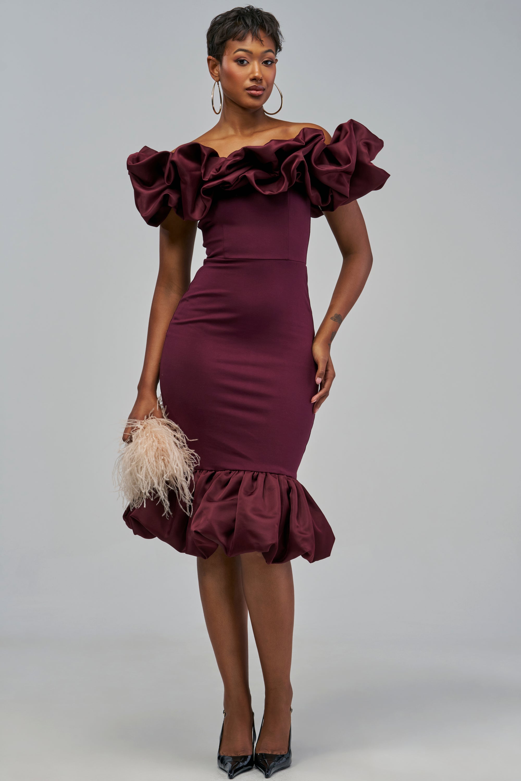 A person wears the COCKTAIL HOUR PUFF SLEEVE MIDI DRESS IN BURGUNDY, an off-the-shoulder ruffle midi dress with a ruffled hem, holding a feathered clutch.