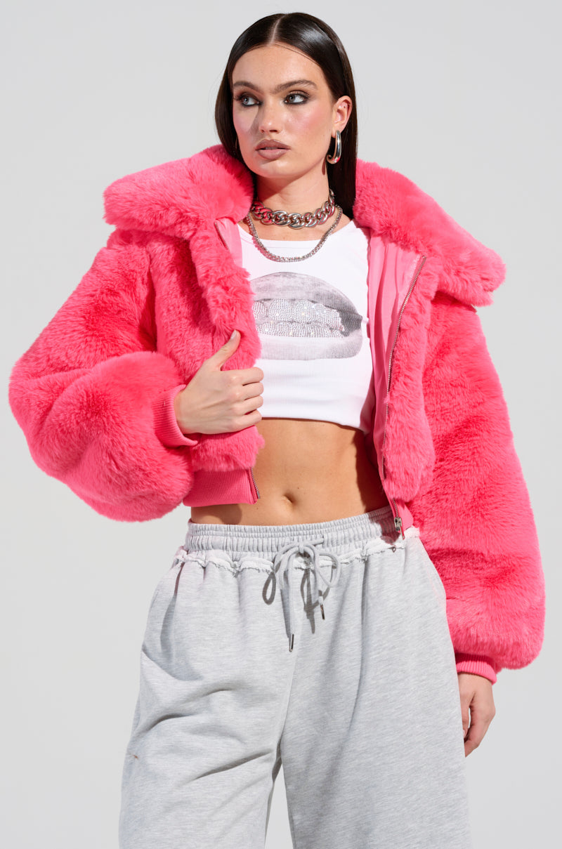 A person poses confidently, wearing the Drew Faux Fur Bomber in hot pink over a cropped white graphic tee. They are also sporting light gray sweatpants, their hands resting in the jacket pockets, perfectly showcasing casual style with a bold pop of color.