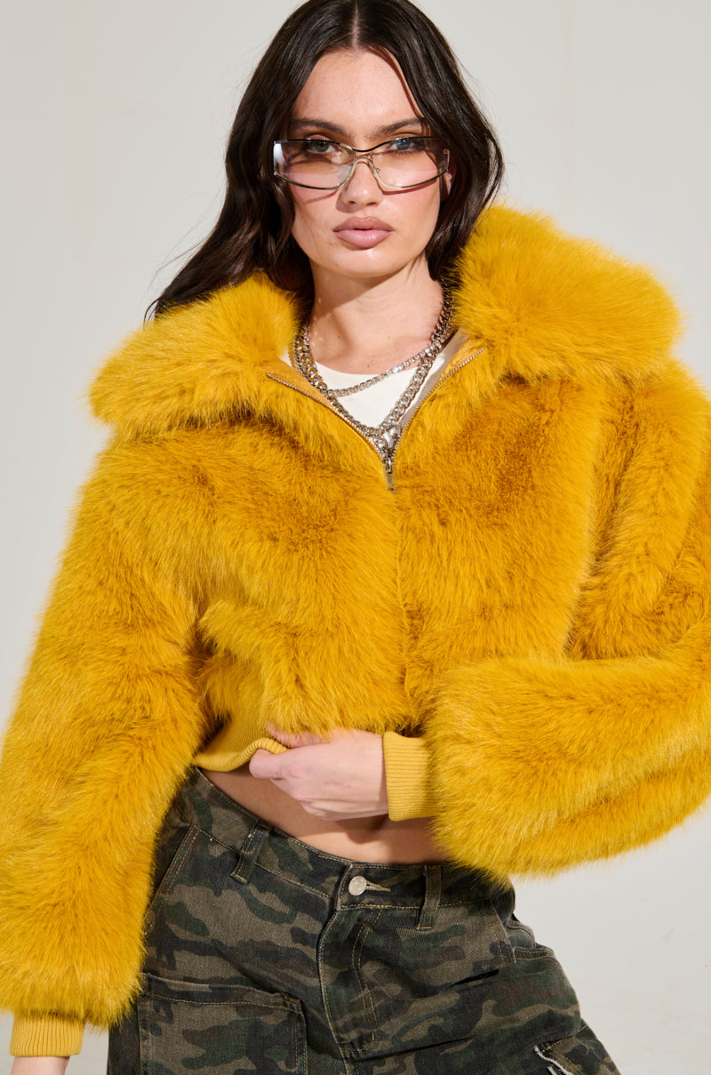 A person confidently poses in the DREW FAUX FUR BOMBER IN YELLOW, a vibrant piece with a cropped silhouette, paired with camouflage pants. They have long dark hair and oversized glasses, adding layered necklaces for extra flair. The neutral background perfectly highlights their bold outfit. 0