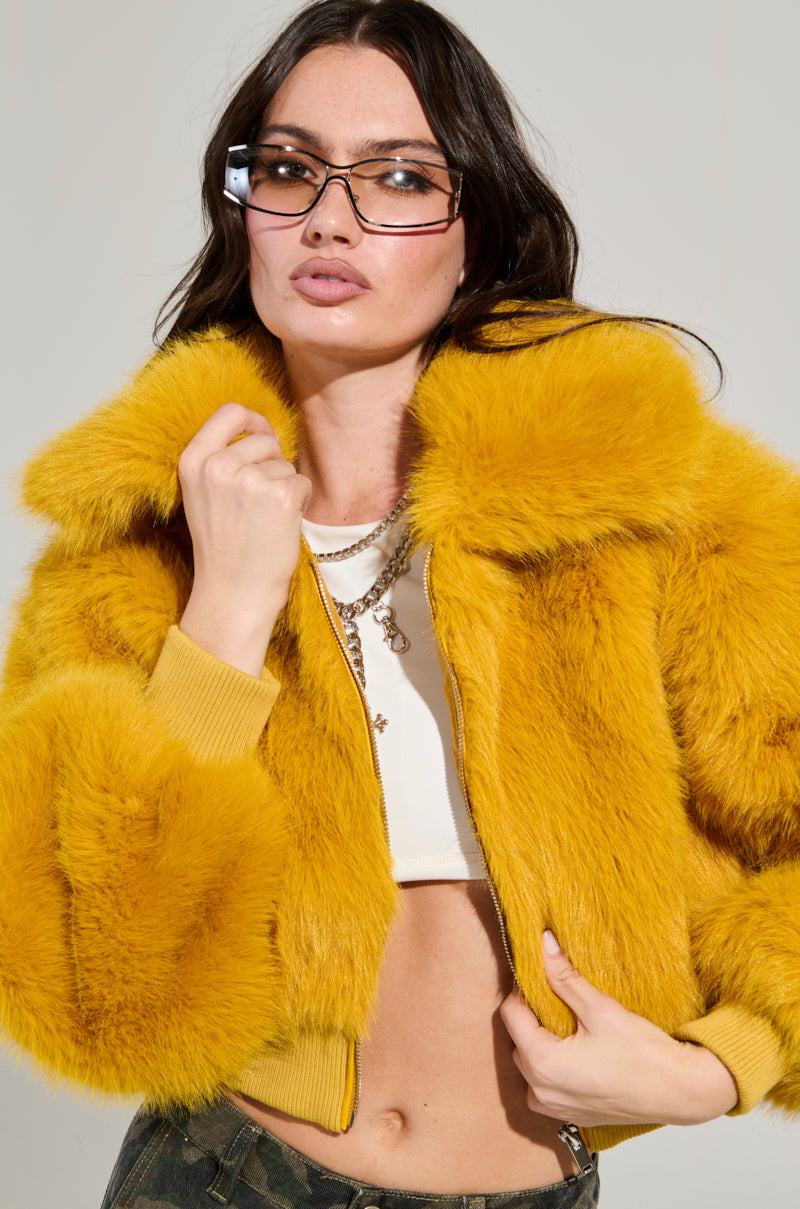 A person confidently poses in the DREW Faux Fur Bomber in Yellow and clear glasses. They sport long dark hair, shown from the waist up in a cropped silhouette with a white top and camouflage pants against a plain background. 2