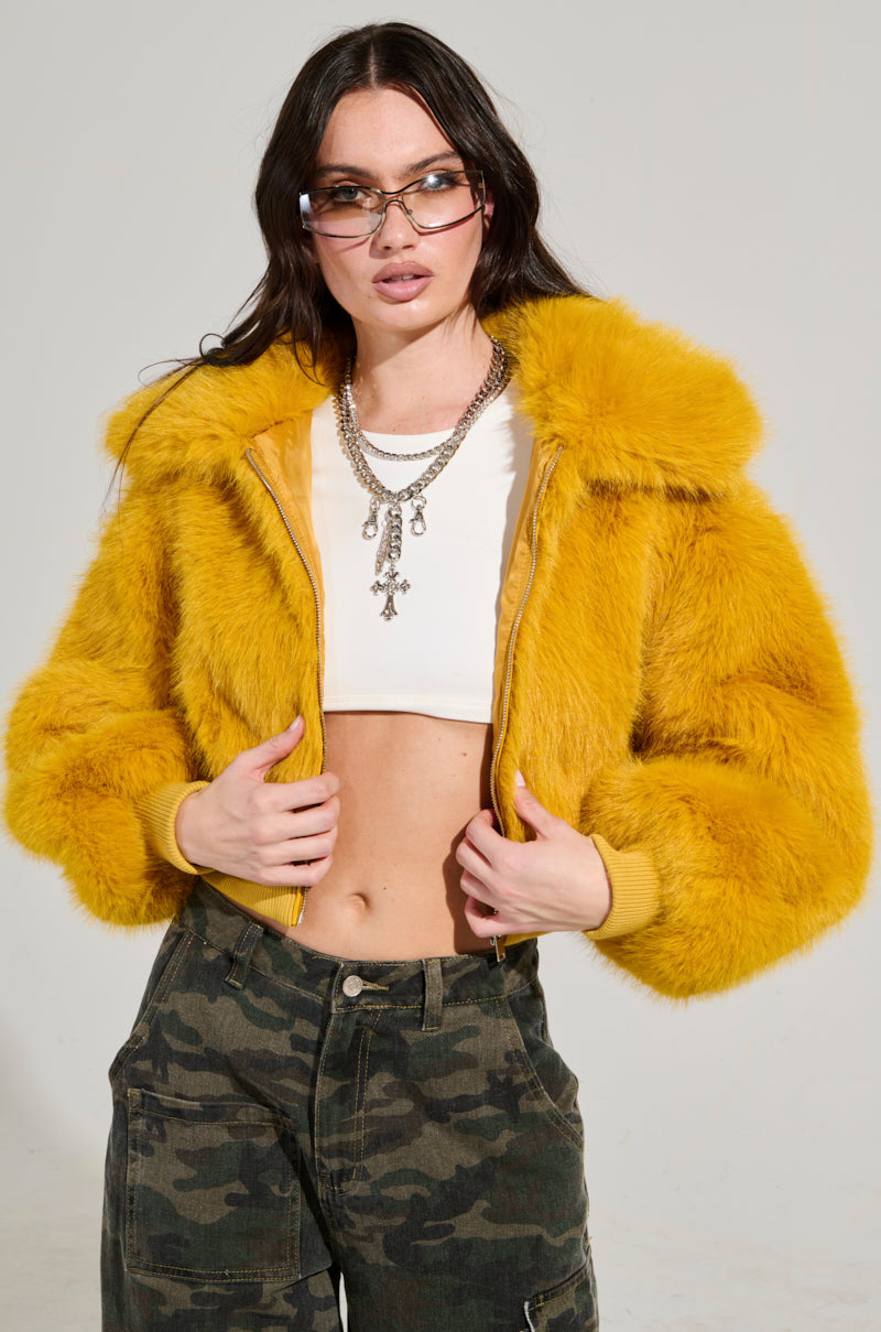 A person showcases the striking DREW FAUX FUR BOMBER IN YELLOW paired with a white cropped top and camo pants. Their long dark hair flows, perfectly framed by sunglasses and layered necklaces against a simple light background. 3