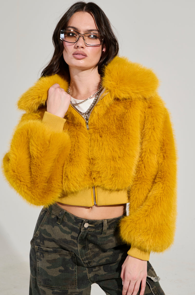 A person confidently poses in the Drew Faux Fur Bomber in Yellow paired with camo pants. They showcase long brown hair, sunglasses, and layered necklaces. The cropped silhouette set against a plain, light-colored background accentuates their bold fashion statement. 5