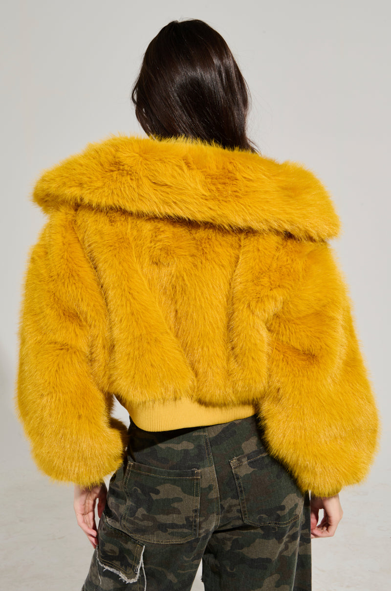 Facing away, a person with long hair showcases the DREW FAUX FUR BOMBER IN YELLOW—a cropped, fluffy jacket with a large collar—perfectly paired with dark camouflage pants. The background is plain and light-colored. 7