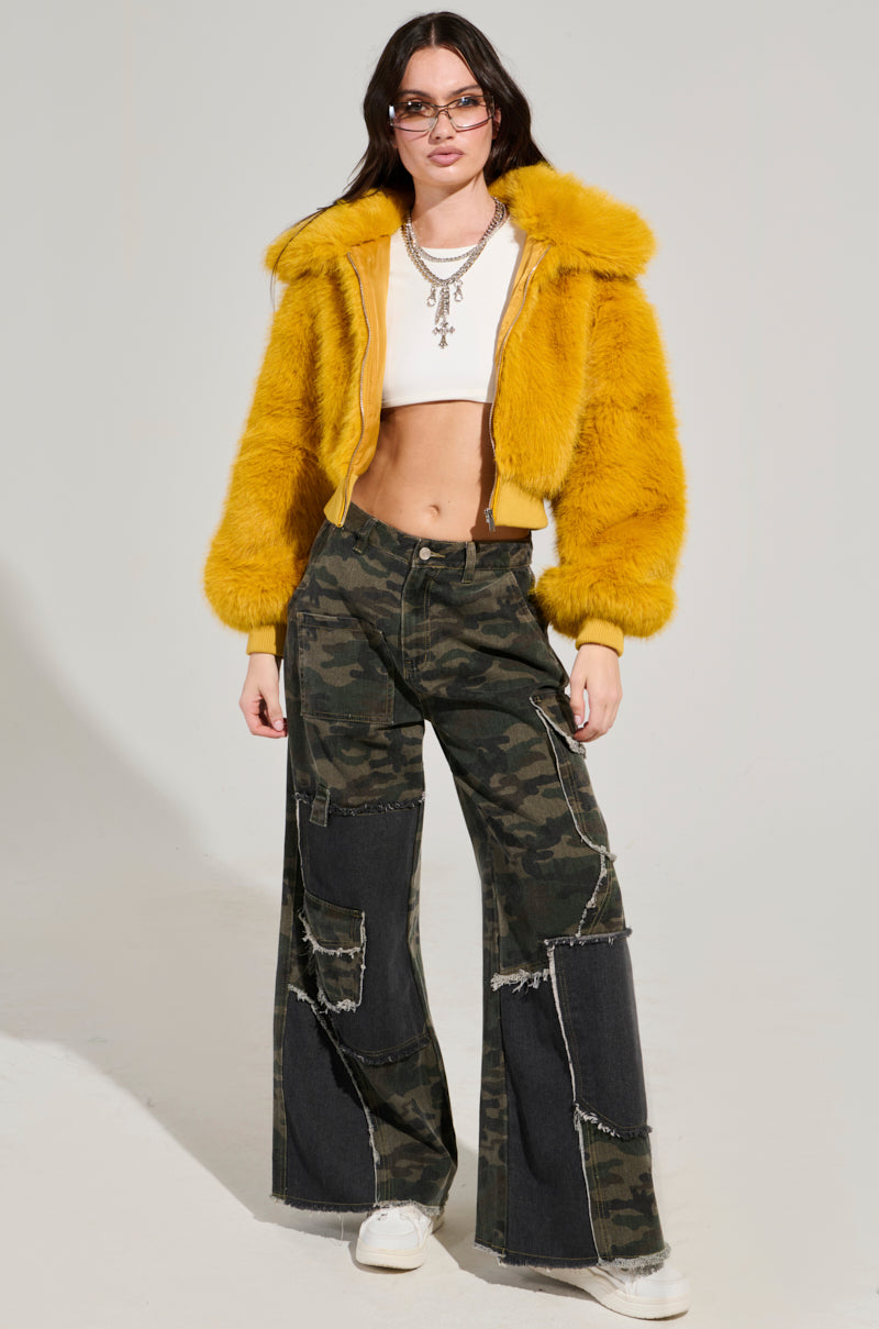A person confidently stands dressed in the DREW Faux Fur Bomber in Yellow, paired with a white crop top and camouflage patchwork pants. They have long, dark hair and sunglasses, accessorized with layered necklaces and white sneakers. 8