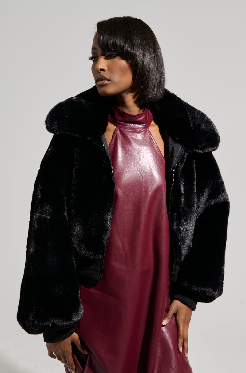A woman poses in a Drew faux fur bomber with rib trim in black over a burgundy leather halter dress. She has sleek, dark hair and is gazing to the side. The monochrome background highlights her elegant style.