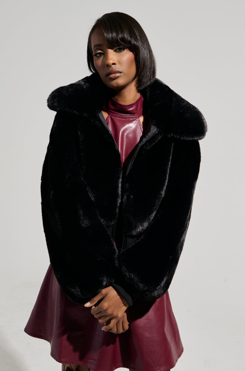 A woman poses in a burgundy leather dress paired with a black Drew faux fur bomber jacket featuring rib trim, set against a light gray background. Her hair is styled straight, and she stands with her hands clasped in front.