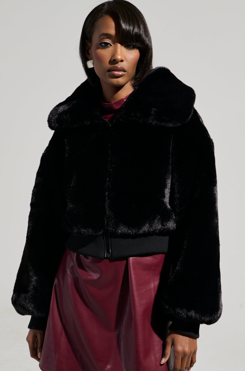 A person wearing the DREW FAUX FUR BOMBER WITH RIB TRIM IN BLACK, featuring a large collar, stands against a plain background. The ensemble includes a maroon top and matching skirt, and the individual has medium-length black hair with a side part.