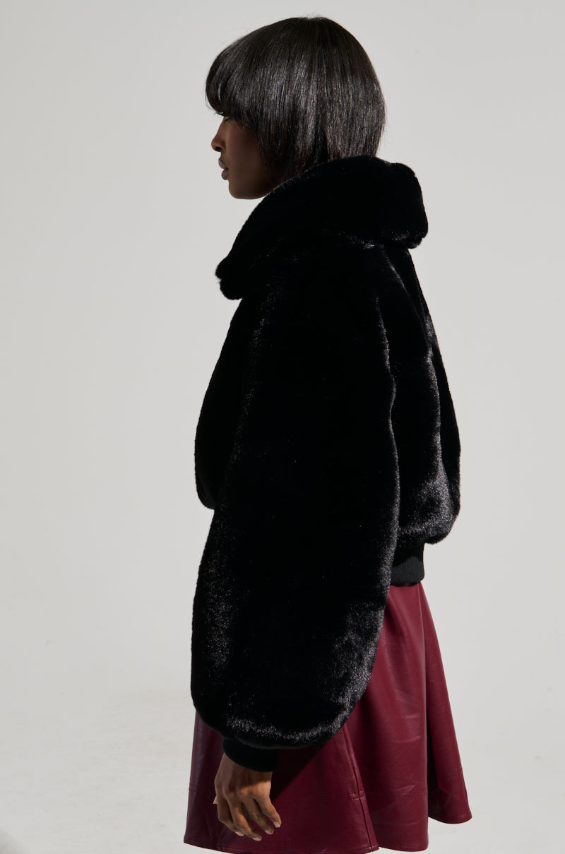 A person with short hair is shown in profile wearing the DREW Faux Fur Bomber with Rib Trim in Black and a maroon skirt, standing against a plain gray background.