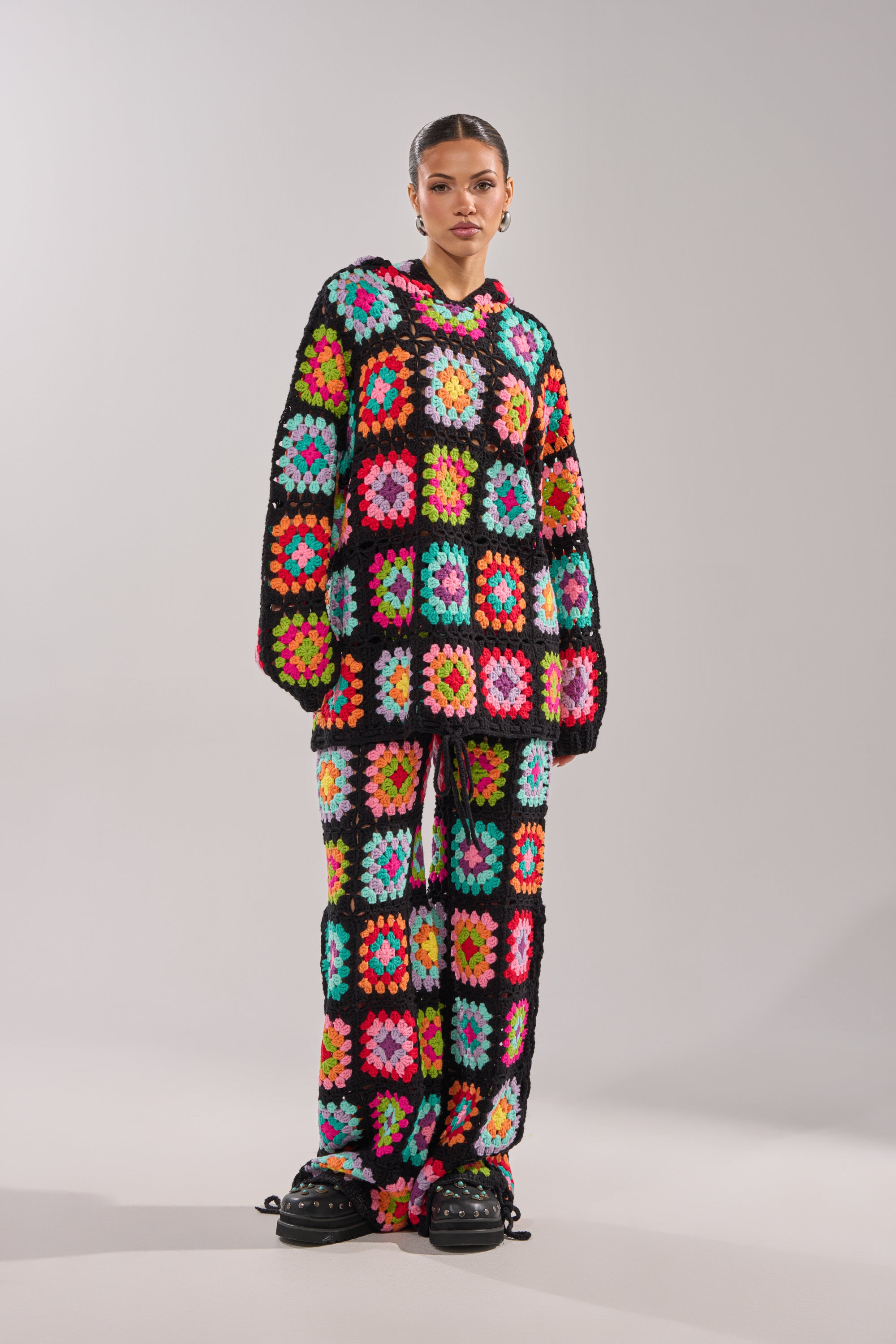 A person wears the FESTIVAL GIRLY OVERSIZED CROCHET SWEATSHIRT IN MULTI, a colorful granny-square patchwork set with black borders, paired with loose pants and chunky black shoes for a vibrant, standout look.