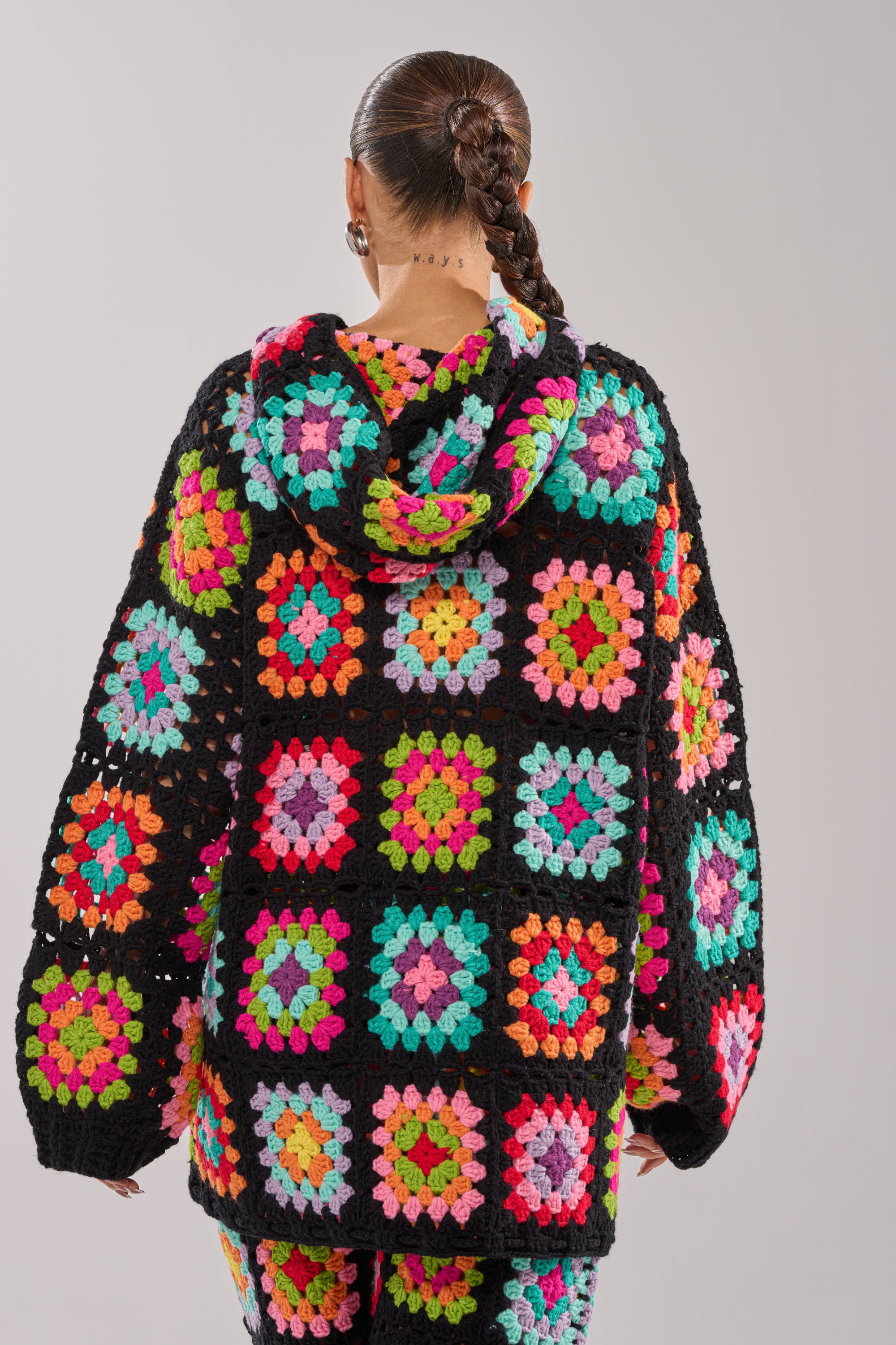 A person with braided hair is seen from behind wearing the FESTIVAL GIRLY OVERSIZED CROCHET SWEATSHIRT IN MULTI, featuring colorful granny square patterns on a black background.